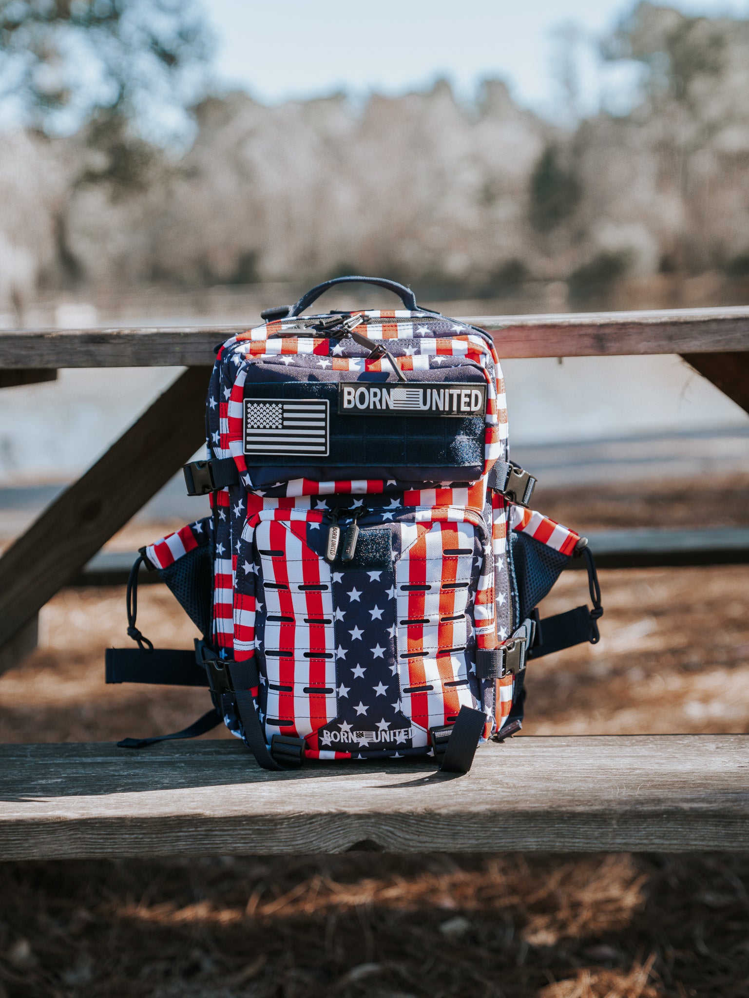 The Ultimate Patriot Special Edition Tac-Pac - 25L by Born United Private Label, with stars and stripes design, sits on a wooden picnic table outdoors, flaunting a U.S. flag patch. Trees and a shimmering lake are visible in the background.