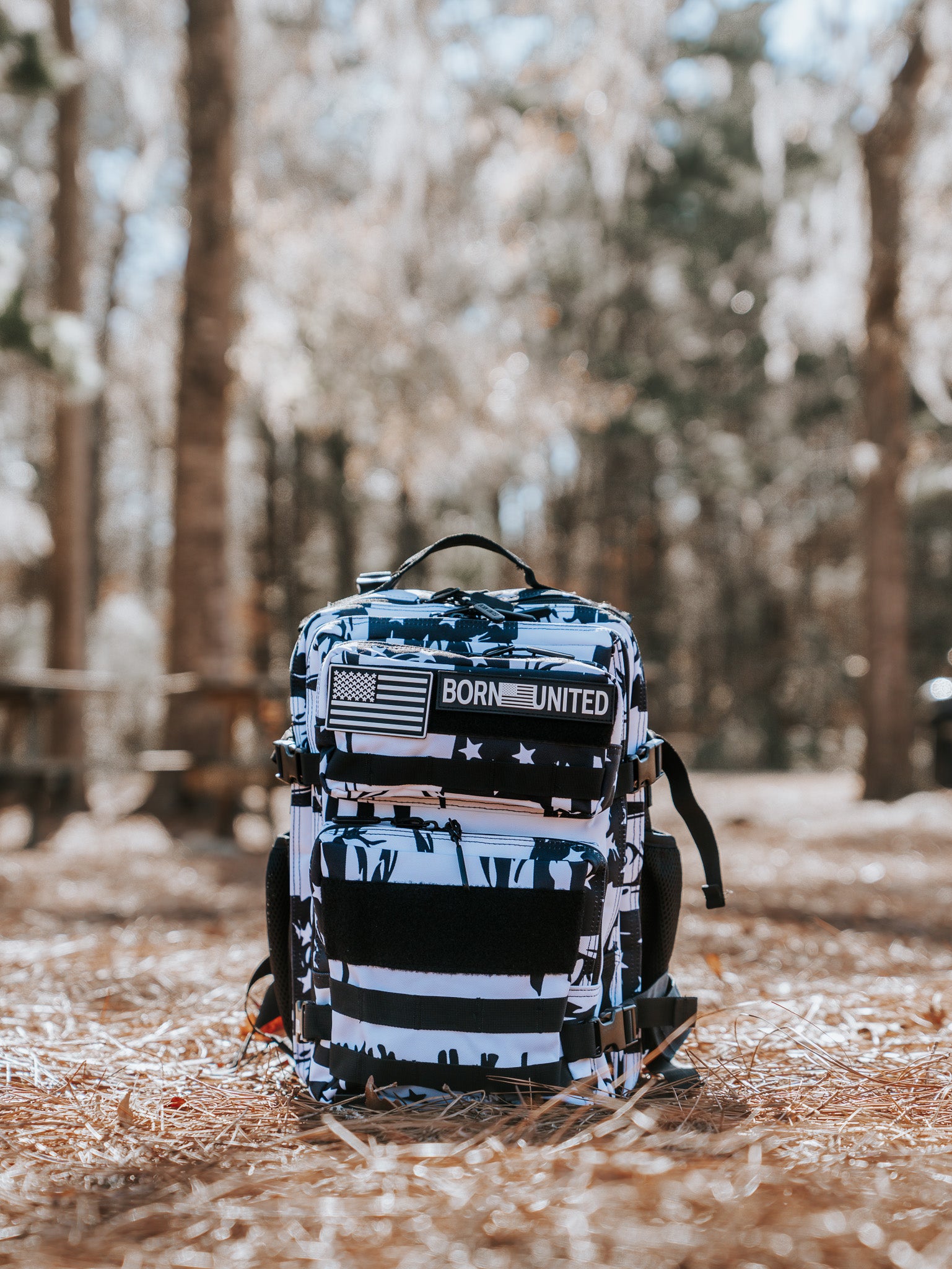 The American Stealth Special Edition Tac-Pac - 25L by Born United Private Label sits proudly on pine needles, featuring a multi-colored design with a Born United patch and American flag. A serene forest scene with blurred trees and picnic tables enriches the backdrop.
