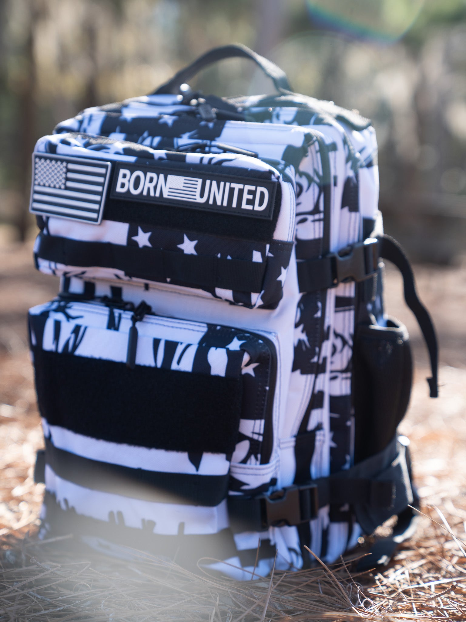 The American Stealth Special Edition Tac-Pac - 25L by Born United Private Label embodies American fashion with its black and white abstract design adorned with stars and a BORN UNITED patch. It rests on dried grass, with blurred trees in the background, echoing stealth clothing essence.