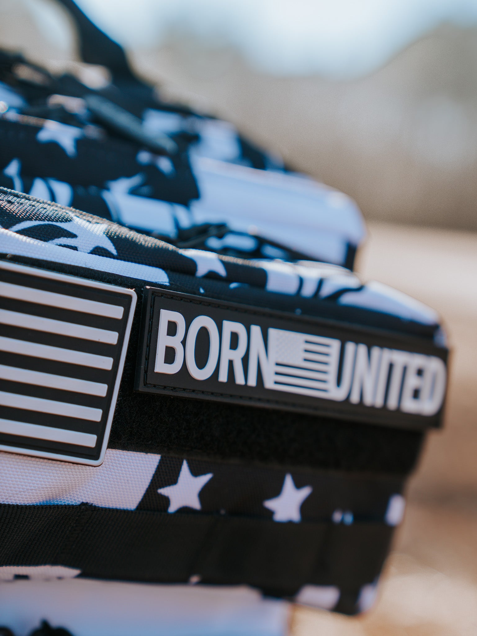 Close-up of the American Stealth Special Edition Tac-Pac - 25L by Born United Private Label. It features a black-and-white American flag pattern with a BORN UNITED patch and an emblem, showcased against a blurred outdoor background.