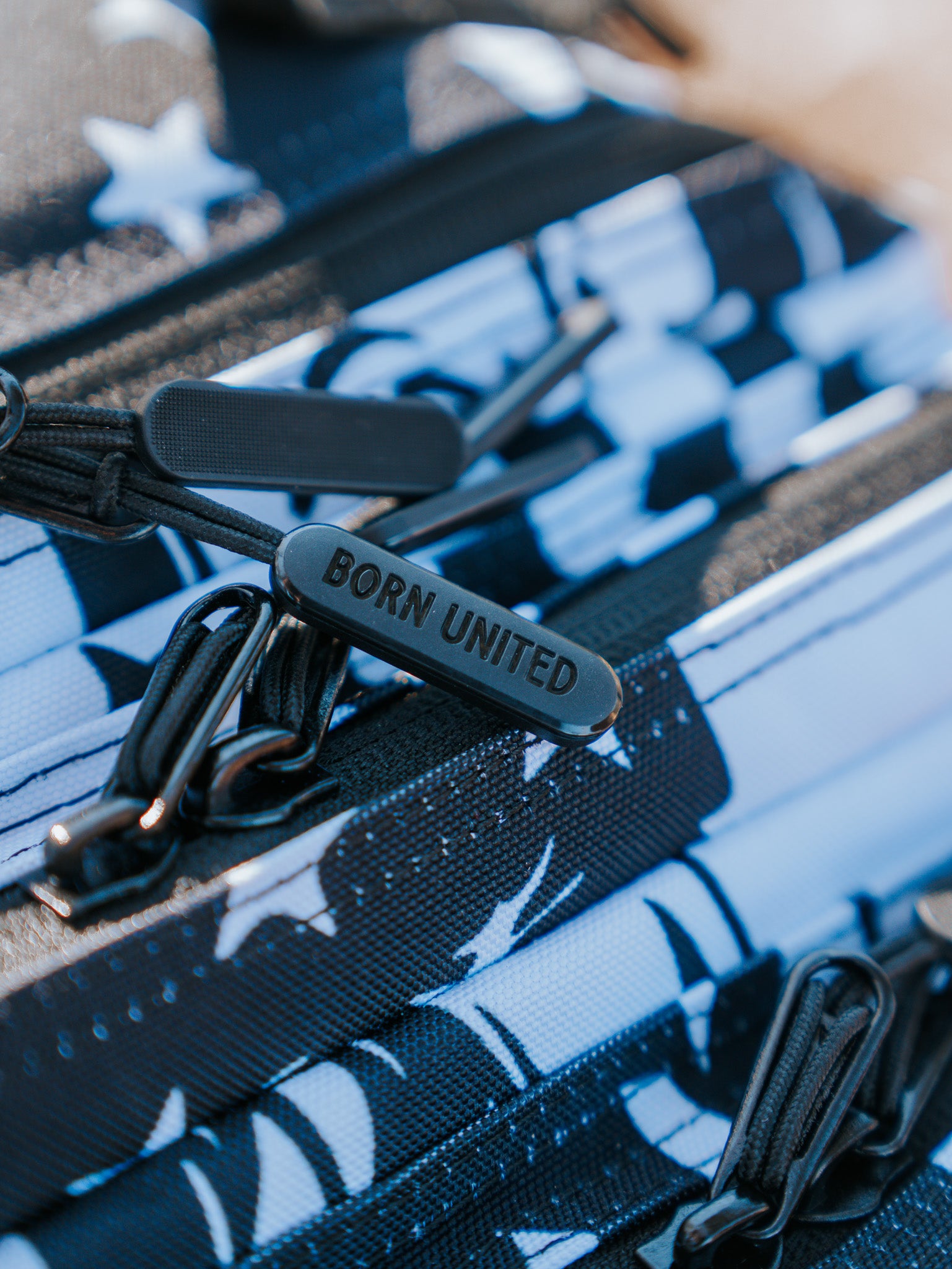 Close-up of a black zipper pull on the American Stealth Special Edition Tac-Pac - 25L by Born United Private Label. This backpack features a bold yet understated geometric pattern in black, white, and blue with star designs and multiple zippers, capturing the essence of modern American fashion.