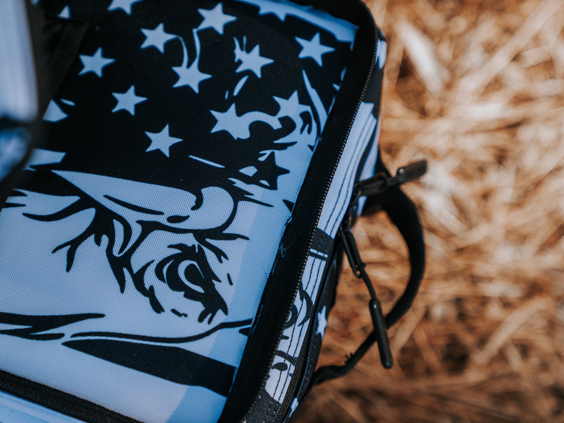 Close-up of an American Stealth Special Edition Tac-Pac - 25L from Born United Private Label, featuring bold black and white graphics with eye and head motifs. Partly zipped, it sits on a straw-like surface under bright lighting, exuding the intriguing world of stealth clothing.