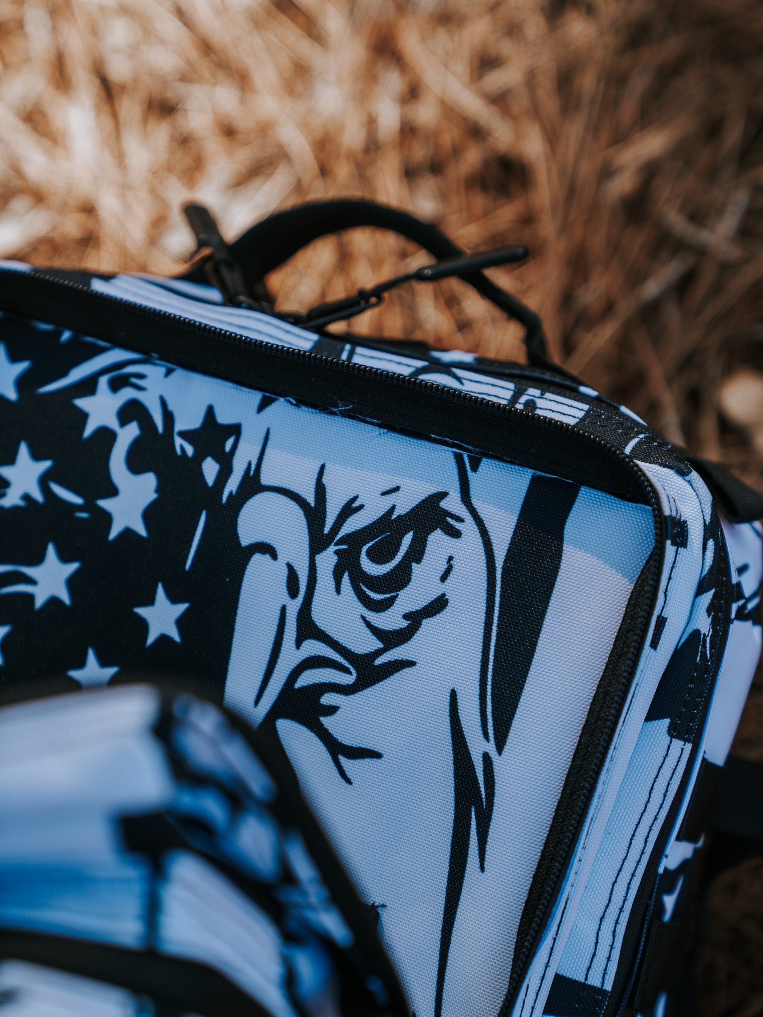 A close-up of the Born United Private Labels American Stealth Special Edition Tac-Pac - 25L features a stylized eagle and stars in black and white, capturing American fashions essence. The dry grass background hints at an outdoor setting where this stealth gear blends into nature seamlessly.