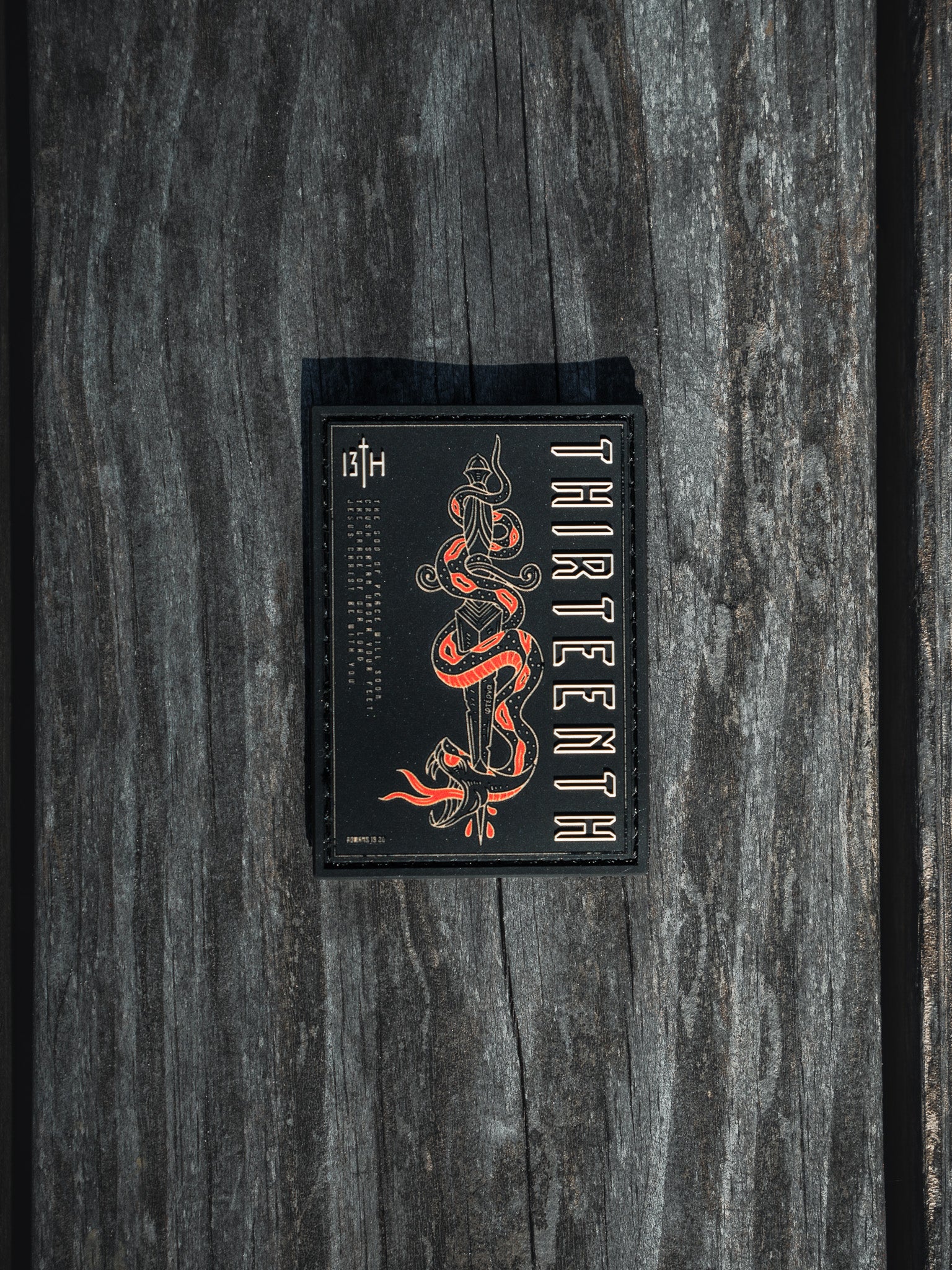 The product, Lucifers Demise PVC Patch by 13th, features a sleek black card with red and white graphics of a coiled snake and the word THIRTEENTH showcased vertically, beautifully set against a textured wooden background.