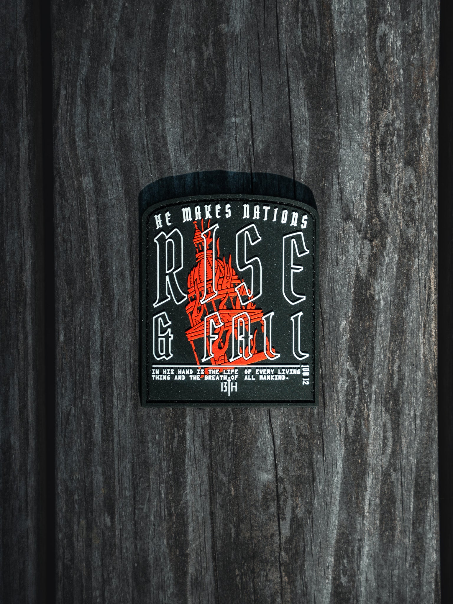 A Rise And Fall PVC Patch on wood features a red sword graphic. The text reads, He makes nations rise & fall. The life hand is in the hands of every living thing and the breath of all mankind. Brand: 13th, Reference: Thirteenth 13:14.