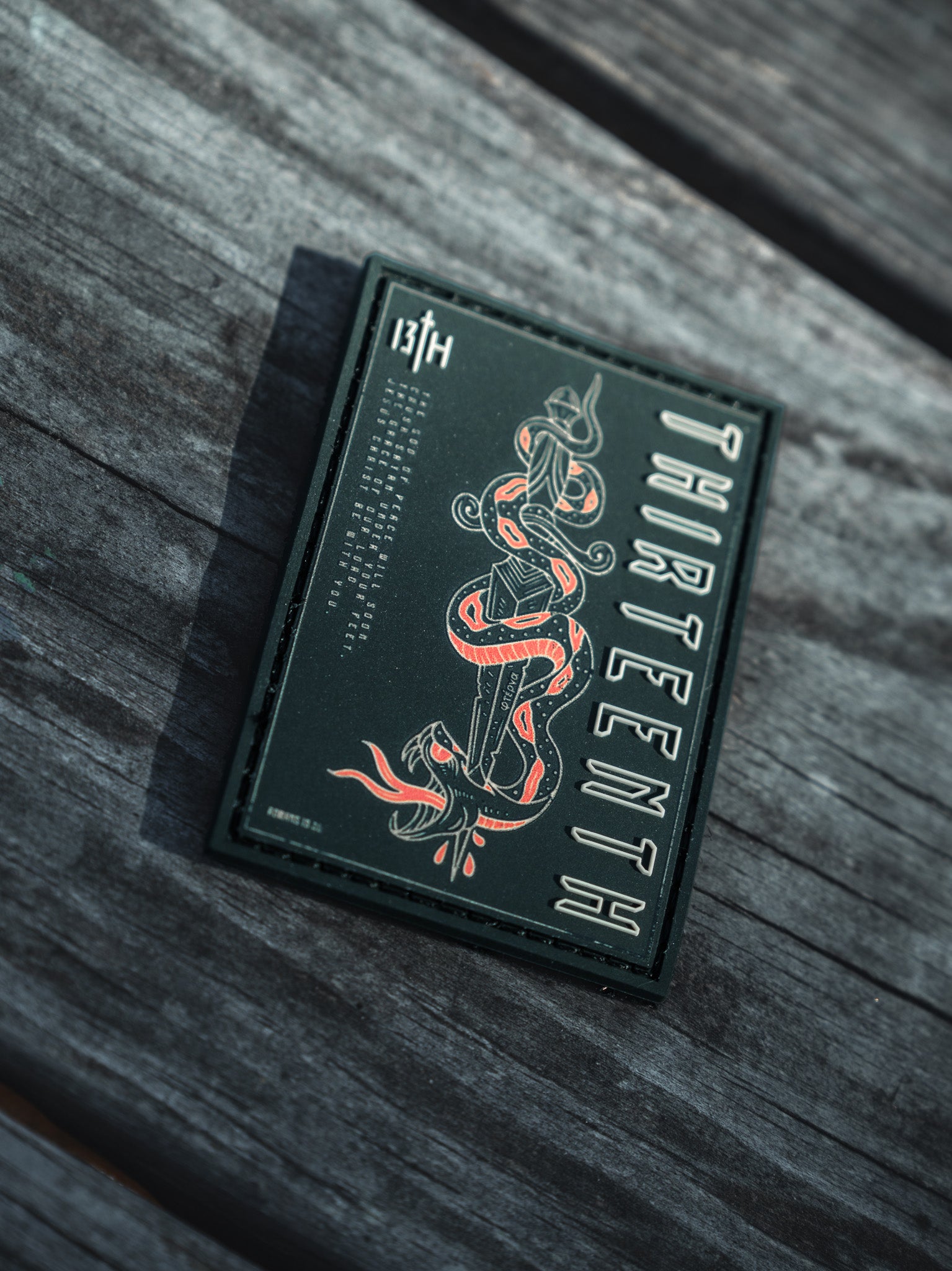 A dark green card showcases the 13th brand vertically and 13TH in the top corner, featuring a striking red and white snake design at its center, known as the Lucifers Demise PVC Patch. Its elegantly displayed on a wooden surface.