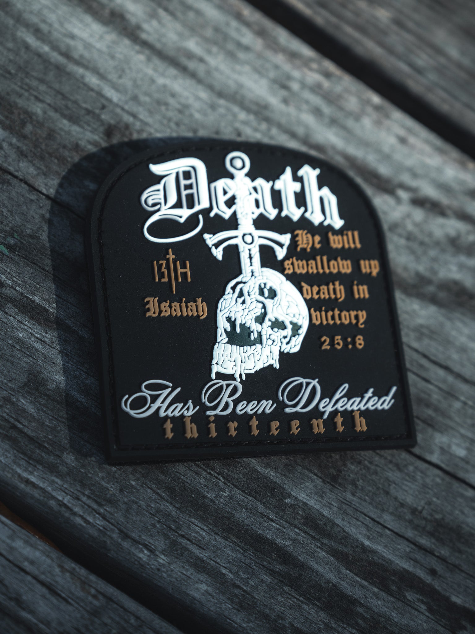 The Deaths Defeat PVC Patch by 13th lies on a wooden surface, showcasing gothic text and a skull. It includes biblical references like Death Has Been Defeated and Isaiah 25:8.