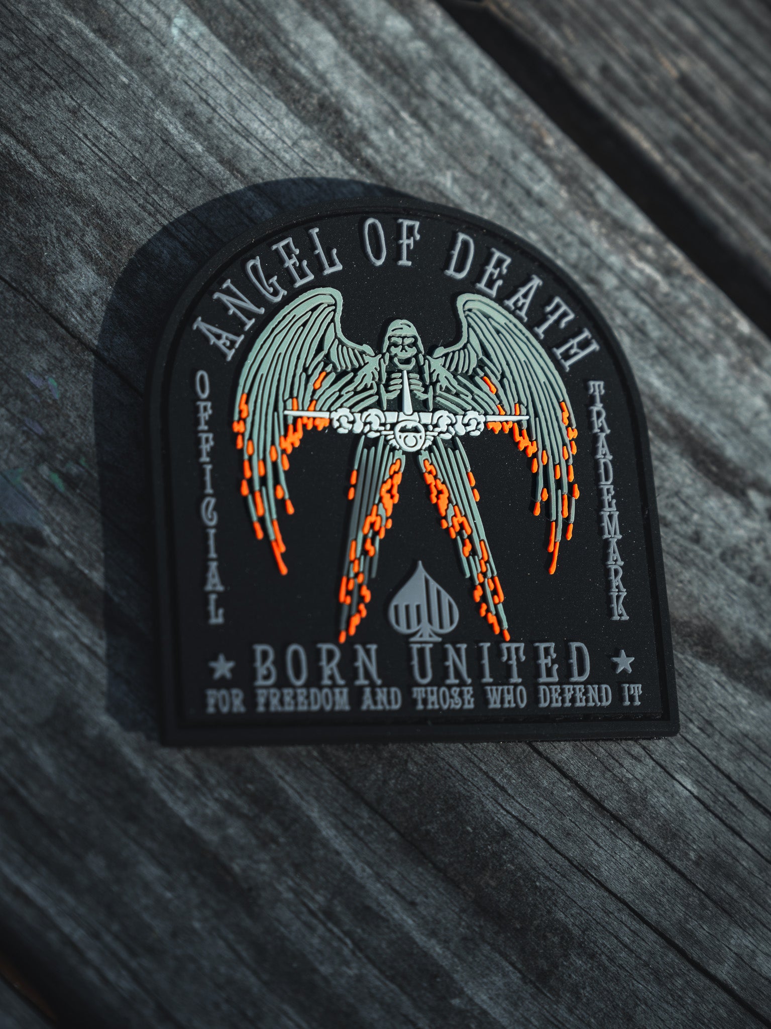 The Angel Of Death PVC Patch by Born United Private Label features an angel with flaming swords and spread wings. Text reads: Angel of Death, Born United, and For Freedom and Those Who Defend It. Displayed on a wooden background, it symbolizes courage.