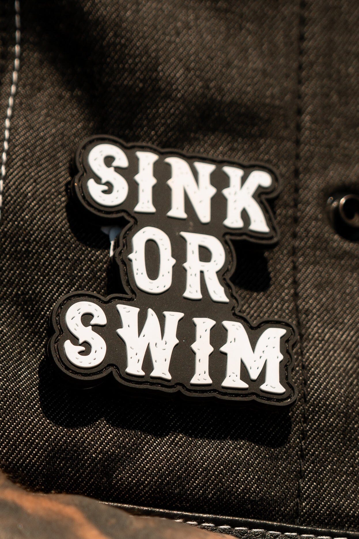 The image showcases a Relentless Betrayal Sink or Swim black and white PVC patch on a denim jacket, boldly capturing the oceans spirit with its distressed lettering.