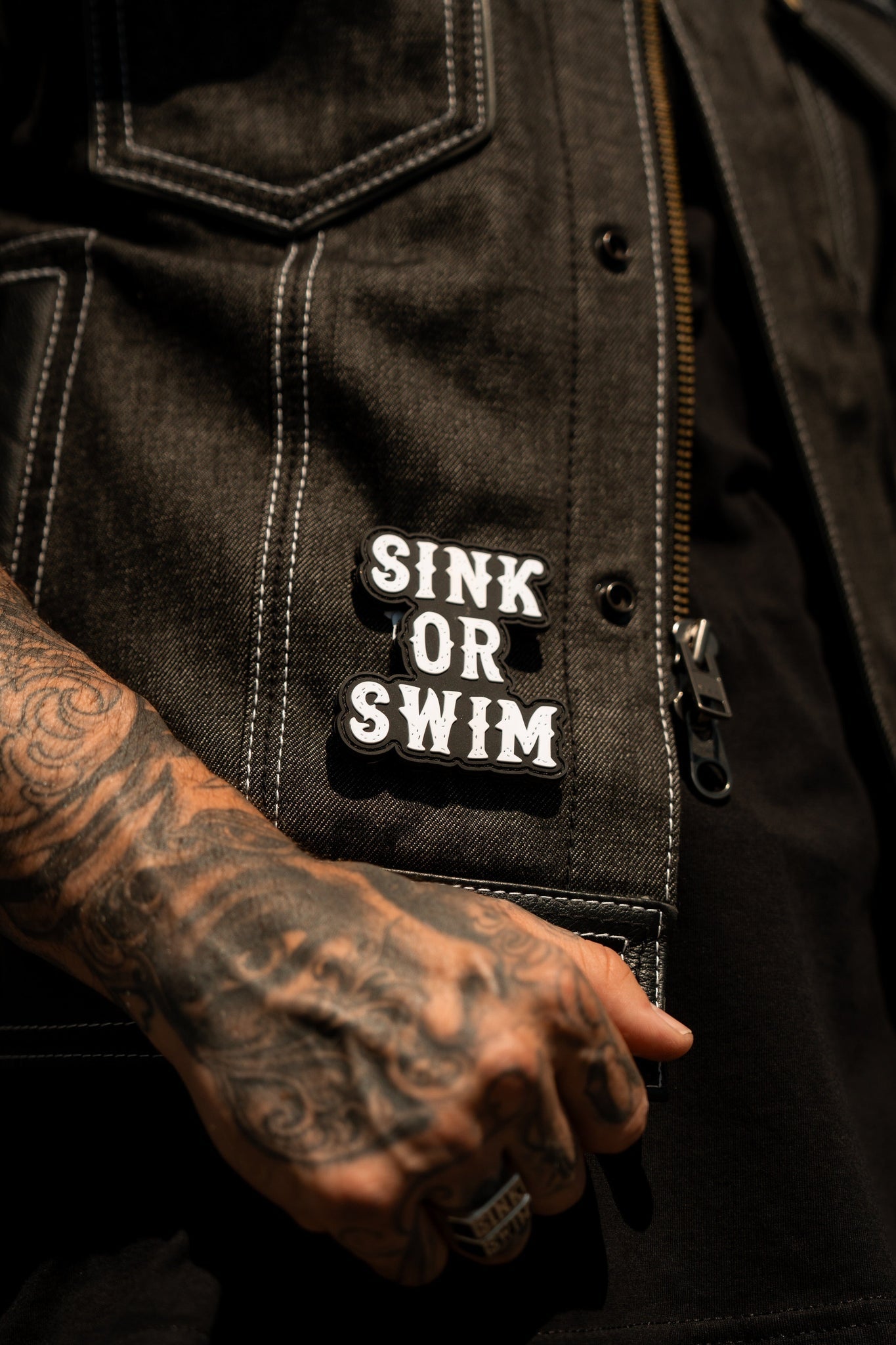 A close-up shows someone wearing a black denim jacket with two SINK OR SWIM pins. Tattooed hands with rings hold the white-stitched edges, revealing an ocean-themed Sink or Swim Black and White PVC Patch by Relentless Betrayal near the zipper.