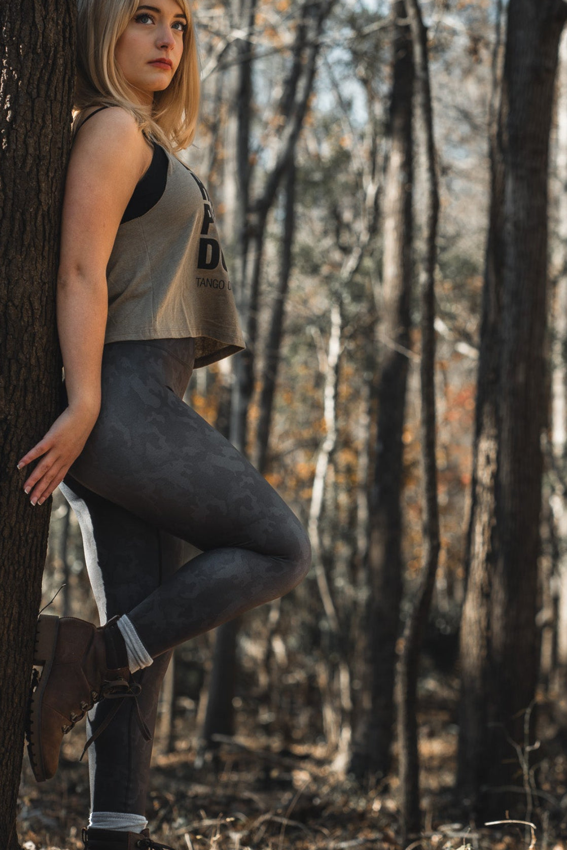 A blonde woman leans against a tree in the forest, wearing Born United Private Labels sleeveless gray top and Midnight Camo Leggings. Her brown boots crunch on dry leaves as sunlight filters through the tall trees.