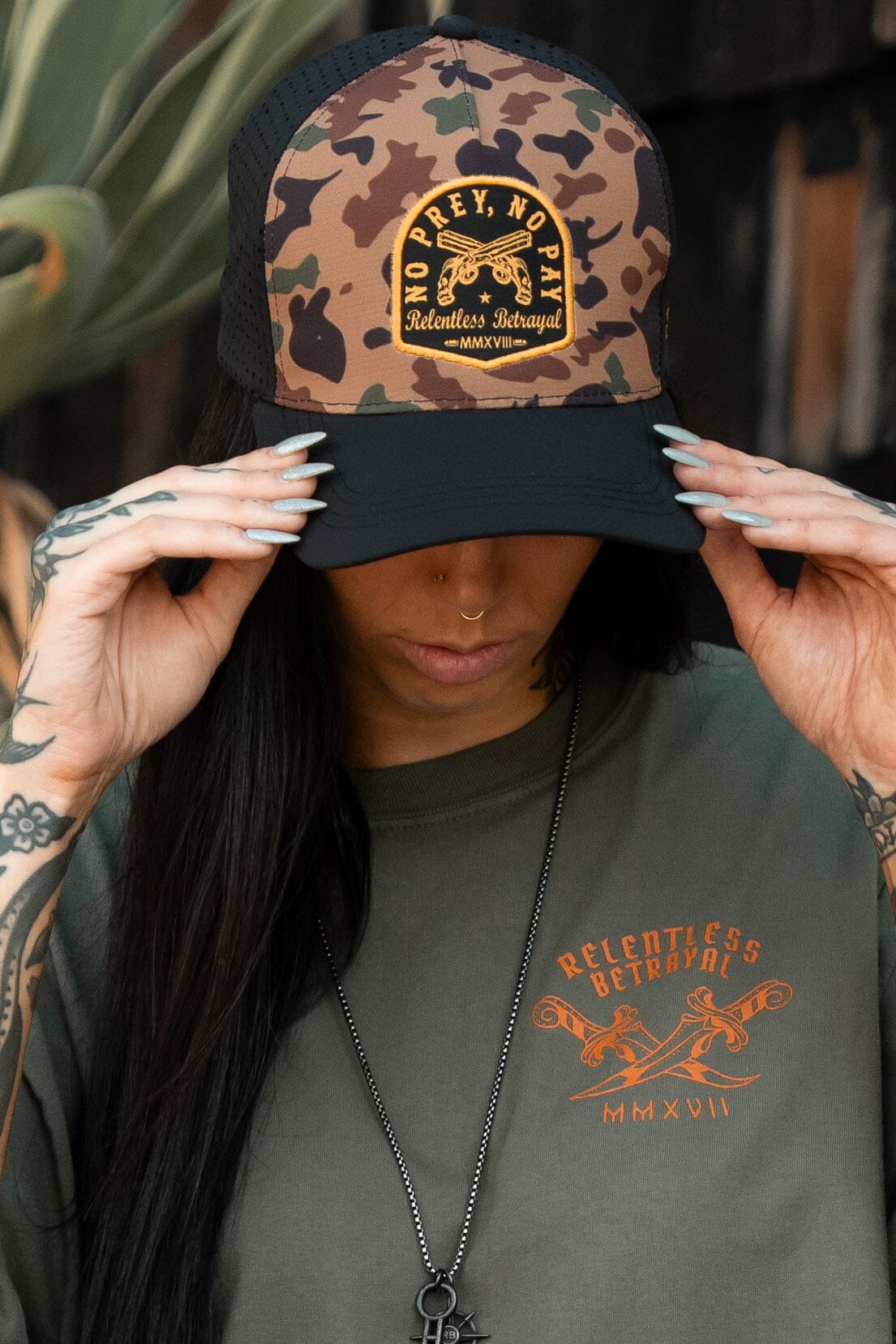 A tattooed, long-haired person adjusts a water-resistant No Prey, No Pay Frog Camo Hat while donning an olive-green Relentless Betrayal shirt with Relentless Betrayal MMXVII. Their manicured nails accentuate their edgy modern pirate lifestyle.