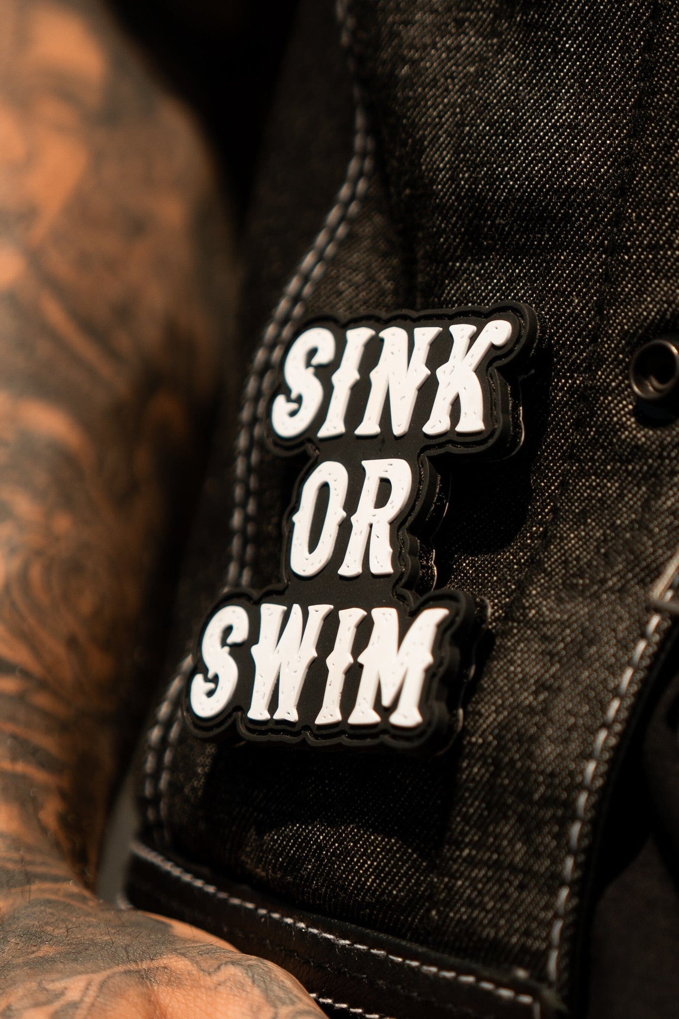 Close-up of Relentless Betrayals denim jacket featuring the Sink or Swim Black and White PVC Patch, enhanced by visible stitching for an edgy look. A tattooed arm beside it evokes a splash of ocean waves, adding personality to the style.