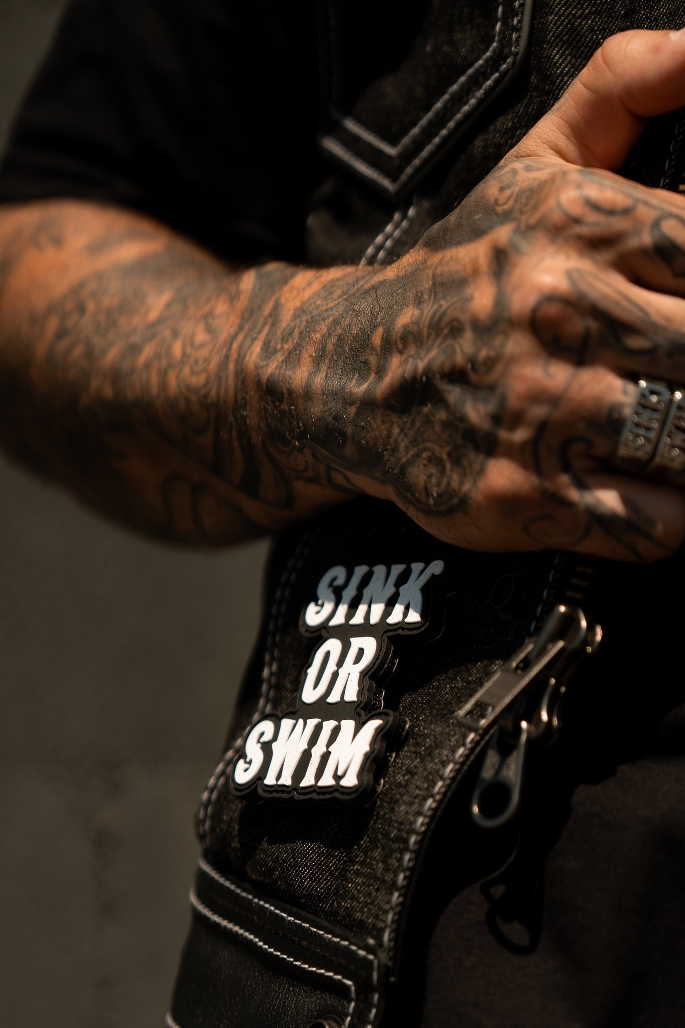 A tattooed hand rests on a black denim jacket featuring Relentless Betrayals striking Sink or Swim Black and White PVC Patch near the zipper. The lighting highlights the detailed tattoos and textured fabric, evoking the oceans intricate beauty.