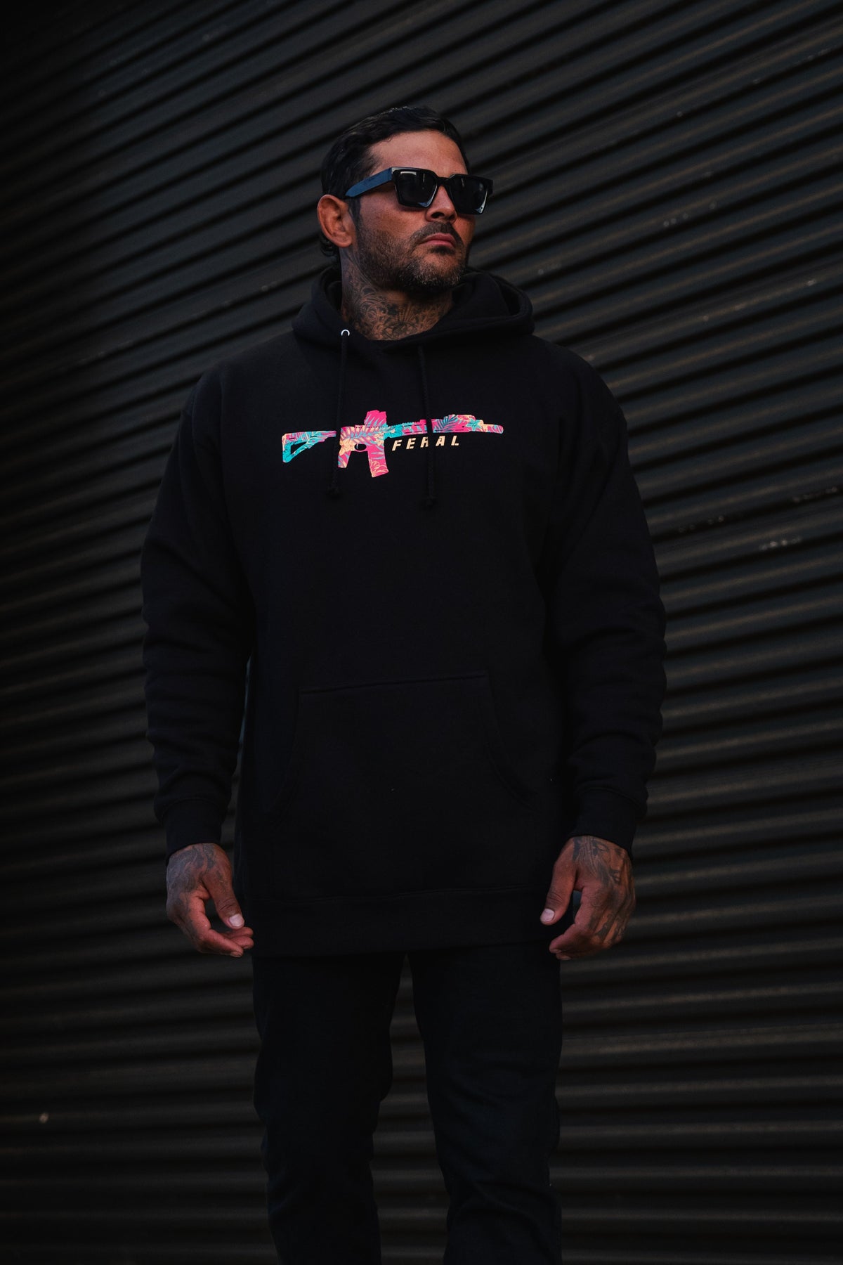 A man wears a black Feral - Premium Hoodie featuring a multicolored rifle graphic and the word FERAL.
