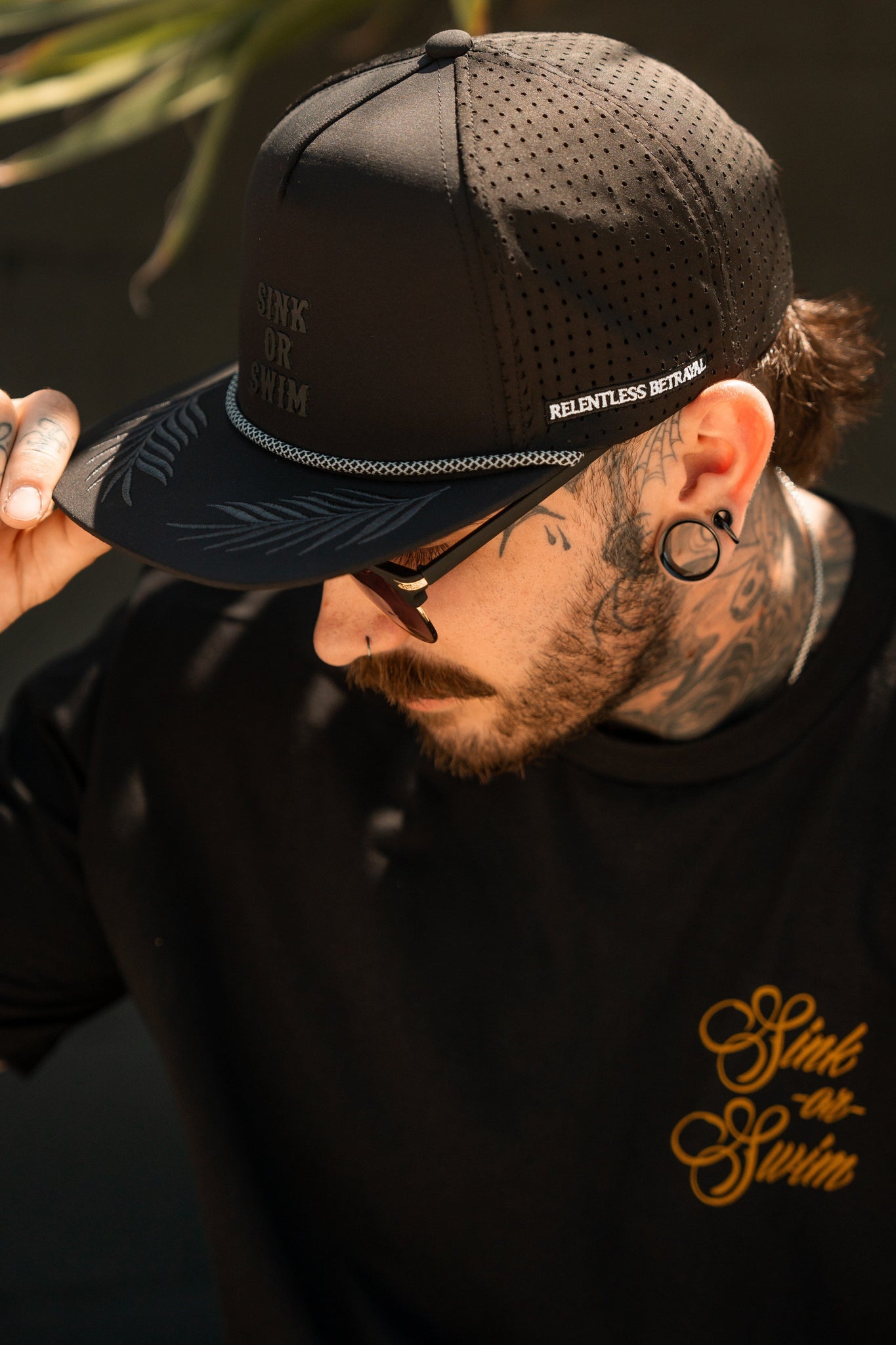 A man wears a Sink or Swim Blackout PVC Snapback by Relentless Betrayal, featuring perforation and water-resistant material, ideal for pirate enthusiasts. He sports a shirt with a gold logo, sunglasses, earrings, and has neck tattoos while outdoors.