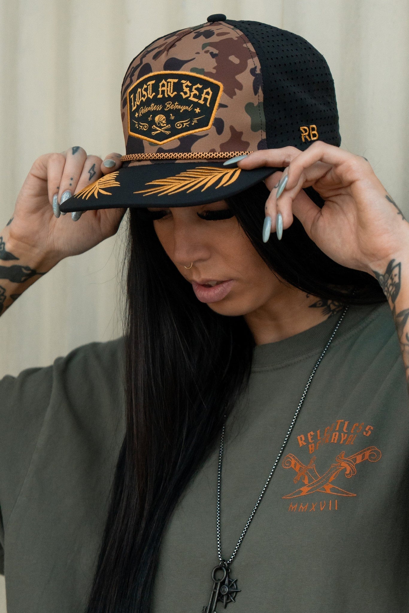 A person with long black hair adjusts a Relentless Betrayal Lost At Sea Frog camo snapback. Theyre in a loose olive green shirt with orange lettering, displaying arm tattoos. A necklace featuring a star pendant completes the look.
