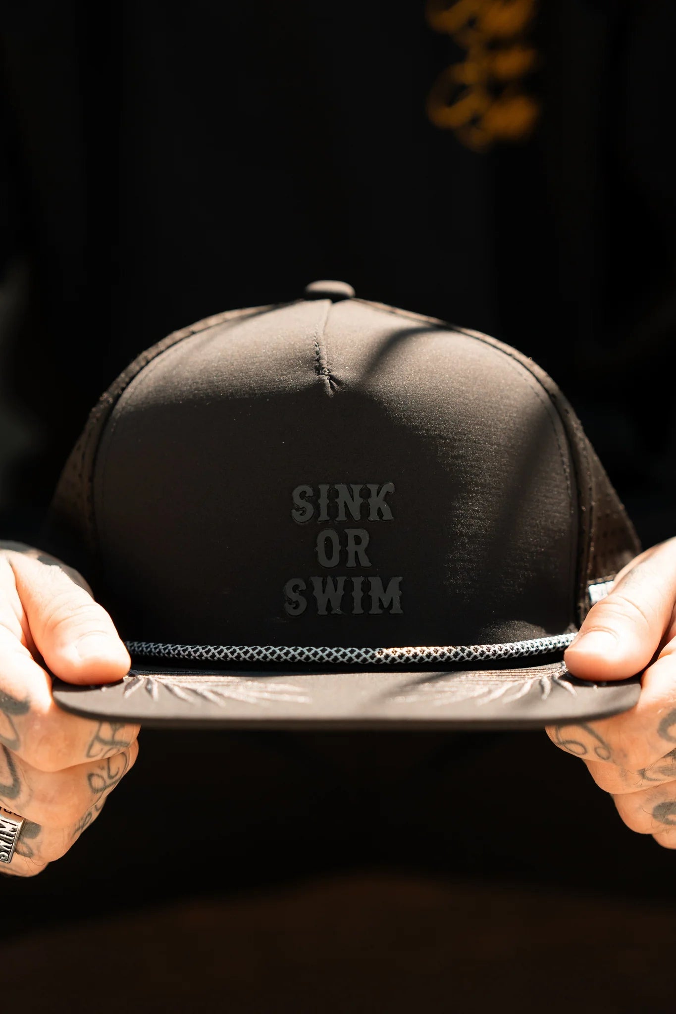 A tattooed individual holds a Relentless Betrayal Sink or Swim Blackout PVC Snapback, with its black and water-resistant design. The dim lighting evokes a mysterious, pirate-like adventure.