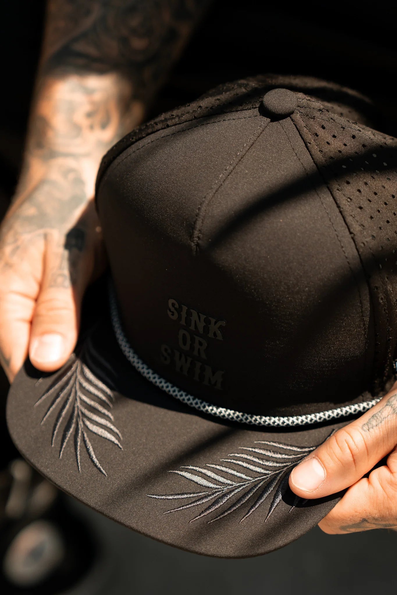 A person with tattooed hands holds the Sink or Swim Blackout PVC Snapback by Relentless Betrayal, showcasing its subtle lettering. This water-resistant cap is decorated with embroidered palm leaves and a braided rope on the brim, capturing the rugged allure of a pirate lifestyle.
