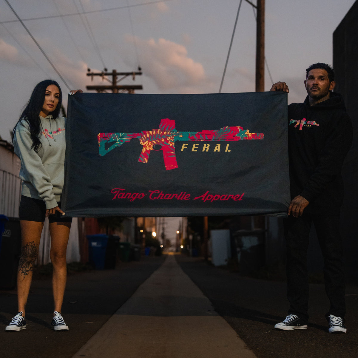 A man and a woman hold a 3x5 Feral - Flag by Tango Charlie Apparel, featuring a red, floral-patterned rifle graphic and the word FERAL.