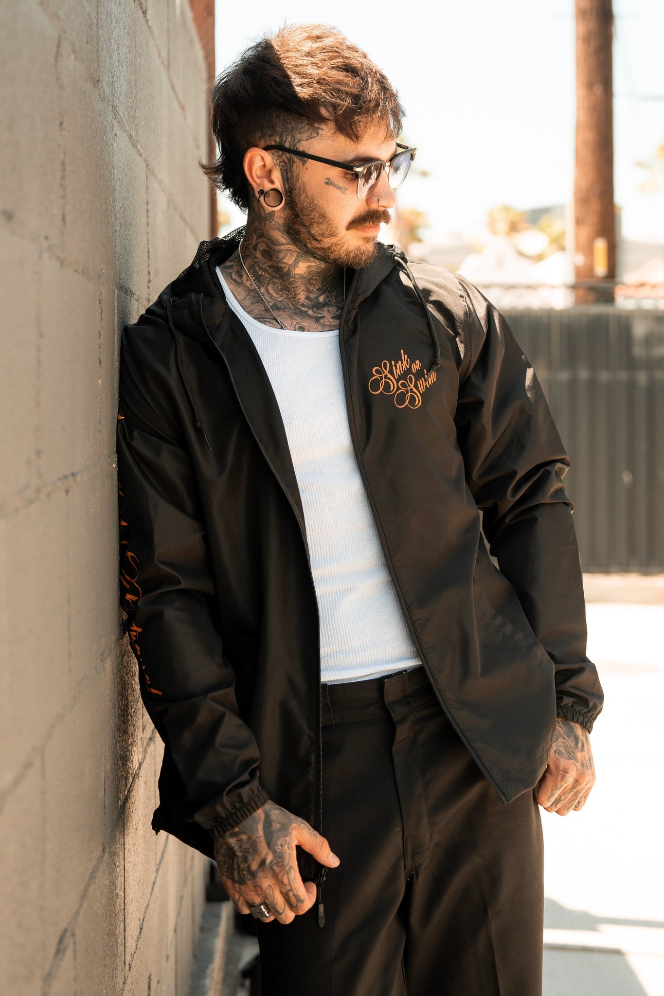 A man with glasses and tattoos leans casually against a wall, wearing a black Relentless Betrayal Sink or Swim full zip jacket with orange embroidery and a white shirt. He has facial piercings and looks downwards as the sunny street extends into the background.