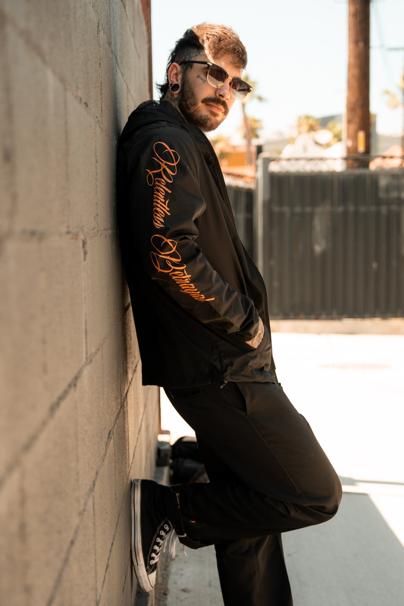 A man leans against a brick wall outdoors, wearing sunglasses and the Sink or Swim Full Zip Jacket by Relentless Betrayal, featuring orange lettering on the sleeve. His black pants and ear piercings complete his look as he gazes sideways, sunlight casting shadows in this modern pirate style.