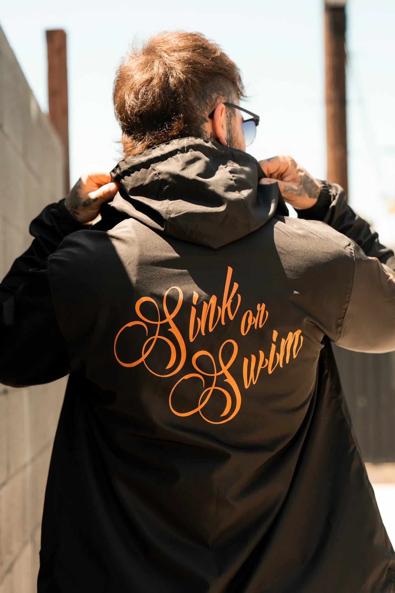 Sporting sunglasses, a person adjusts the hood of their black Relentless Betrayal Sink or Swim Full Zip Jacket, featuring an orange cursive phrase on the back. They stand outdoors, next to a brick wall, embodying a daring pirate lifestyle.