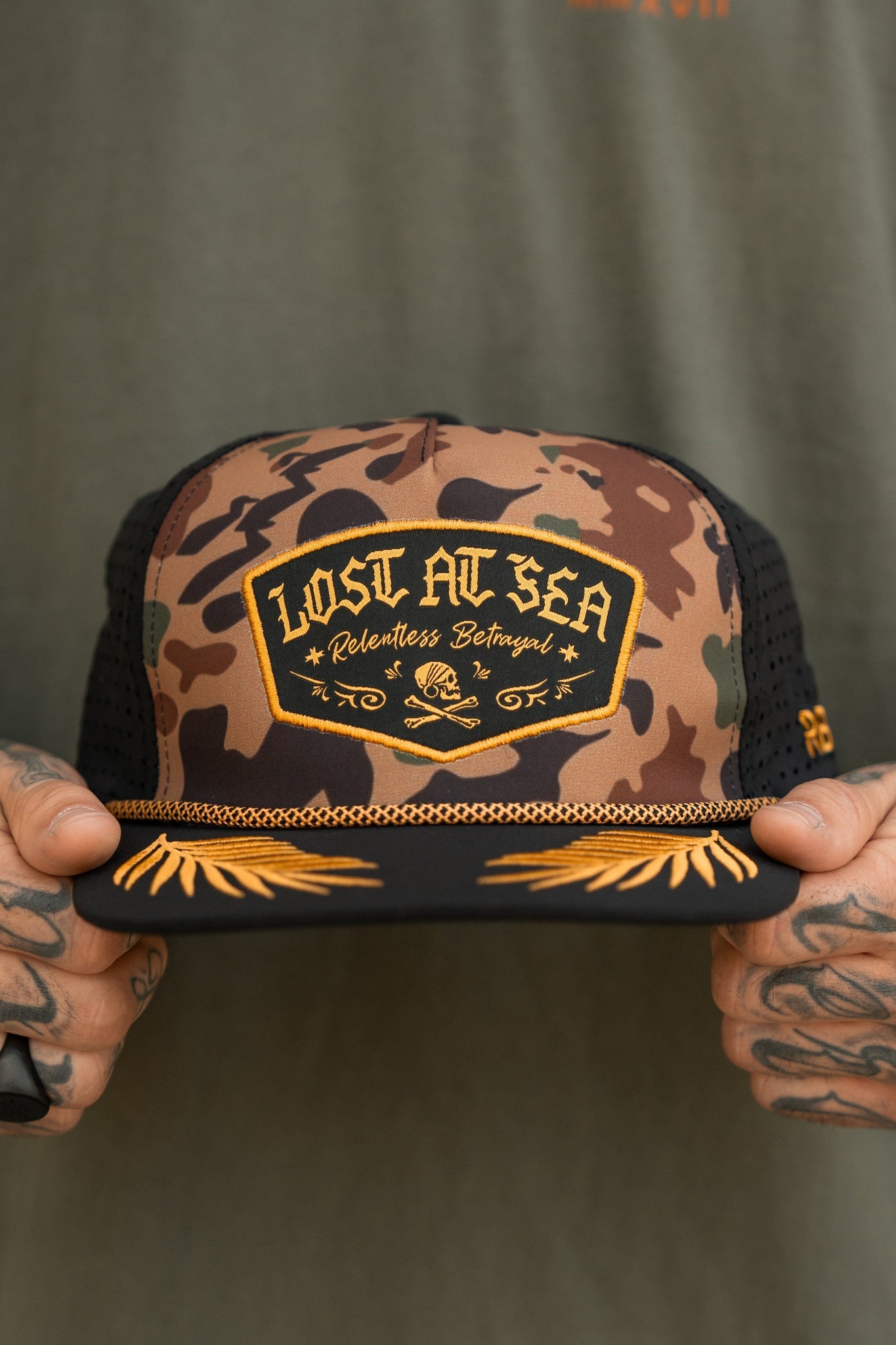 A person holding a water-resistant Lost At Sea Frog Camo Snapback by Relentless Betrayal, featuring gold embroidered skull and crossbones accents, complements their tattoos and green shirt.