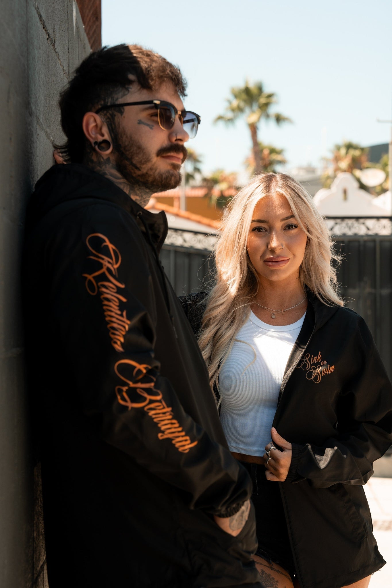 A man and woman showcase their black Sink or Swim Full Zip Jackets by Relentless Betrayal next to a wall. The woman pairs hers with a white top; the man adds sunglasses and tattoos. With palm trees and a fence in the clear skys backdrop, they capture a pirate lifestyle vibe.