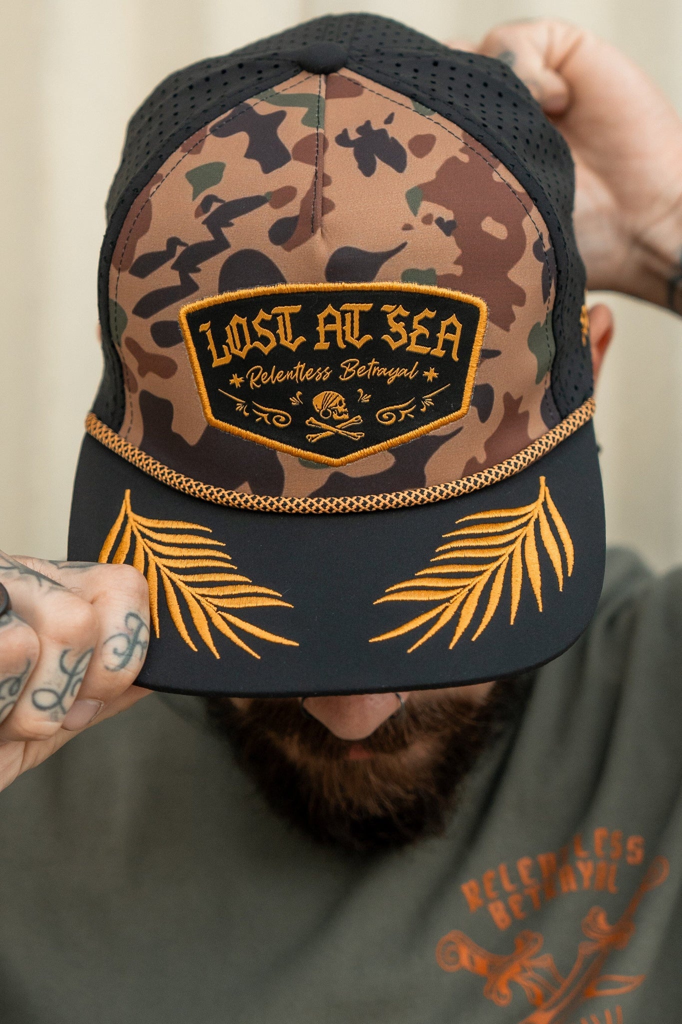 A person adjusts their Lost At Sea Frog Camo snapback by Relentless Betrayal, its patch glinting. The hat, with golden palm leaf embroidery and water resistance, complements their green shirt featuring a matching design.