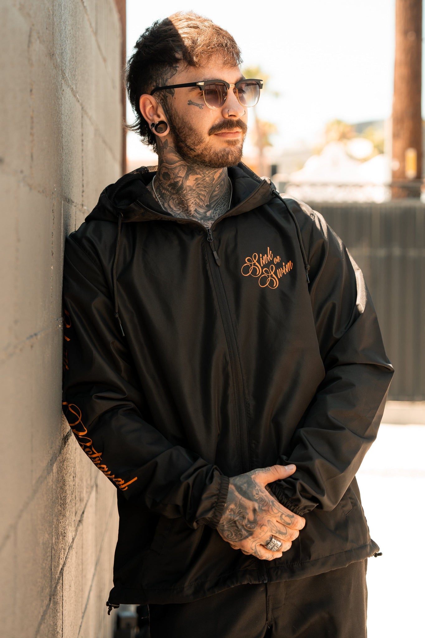 A tattooed person with ear gauges leans against a wall, embodying a pirate lifestyle. They wear shades and a sleek black Sink or Swim Full Zip Jacket by Relentless Betrayal with orange lettering. The outdoor backdrop of a fence and trees adds an adventurous vibe to the scene.