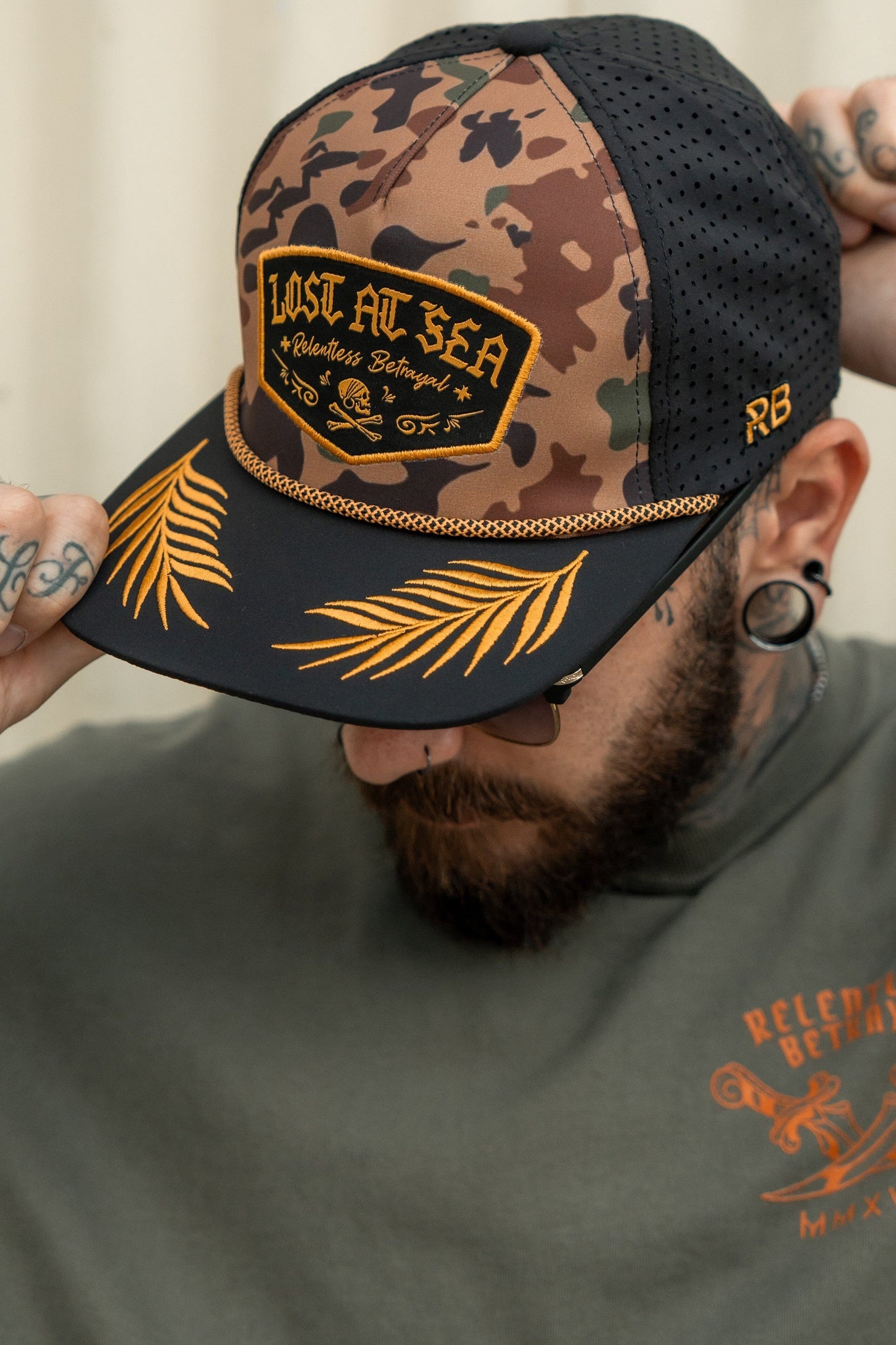 With tattooed hands, someone adjusts their Relentless Betrayal Lost At Sea Frog Camo snapback. The skull design, palm leaf embroidery, RB initials, sunglasses, and gray octopus logo shirt finalize the water-resistant hat’s adventurous look.
