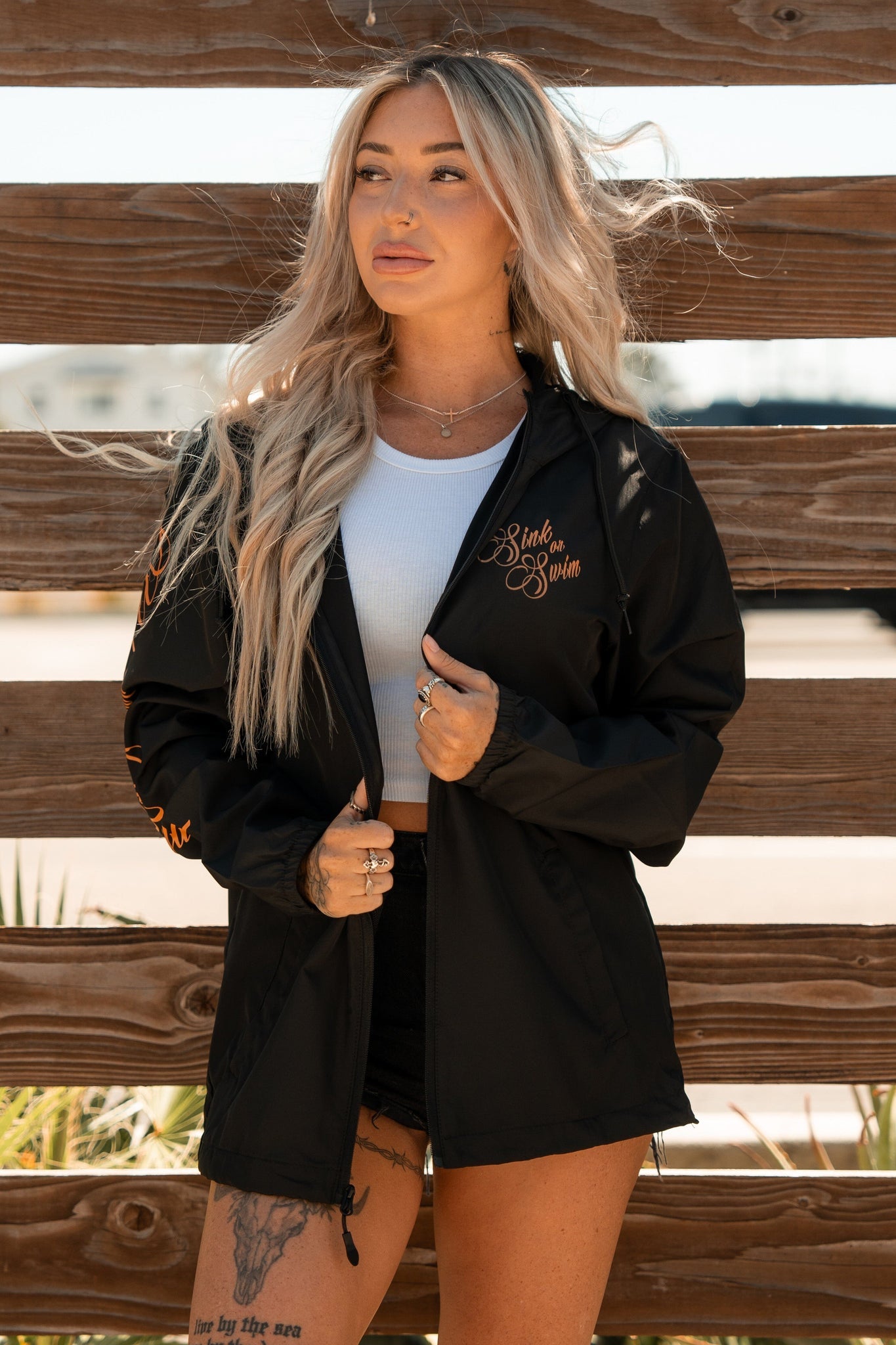 A long-haired blonde woman stands by a wooden fence in the Relentless Betrayal Sink or Swim Full Zip Jacket, over a white crop top and black shorts. Her hands hold the jackets zipper, revealing tattoos on her left thigh, giving her an edgy pirate aura.