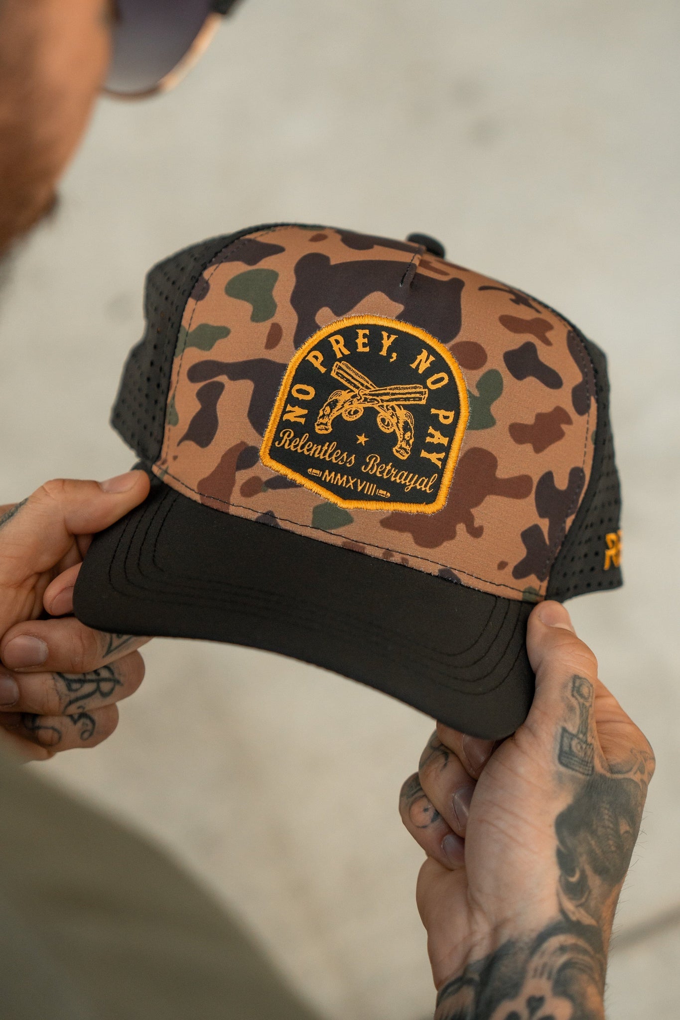 A person with tattooed hands holds the No Prey, No Pay Frog Camo Hat by Relentless Betrayal, a water-resistant snapback with a black bill. The cap has a patch saying No Prey, No Pay and Relentless Betrayal MMXXVII, plus a skeleton and ship wheel graphic symbolizing the pirate lifestyle.