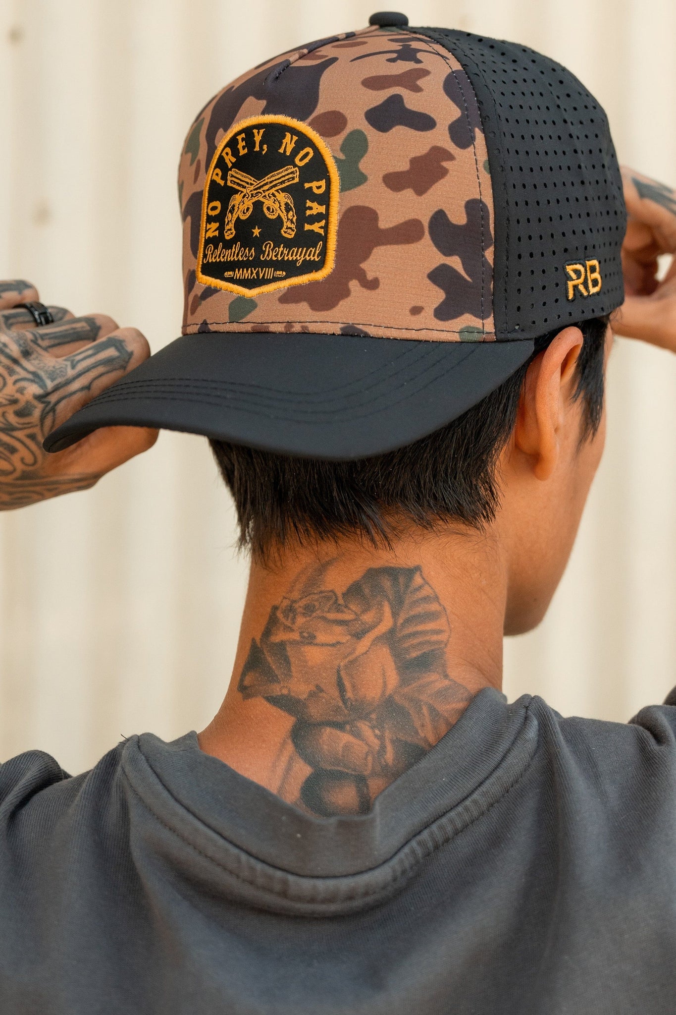 A tattooed person adjusts a water-resistant No Prey, No Pay Frog Camo Hat by Relentless Betrayal, featuring NO PREY, NO PAY Subtleties Betrayal MMWVIII and RB initials. With its black knit back, it embodies the rugged pirate essence. Theyre wearing a dark shirt.
