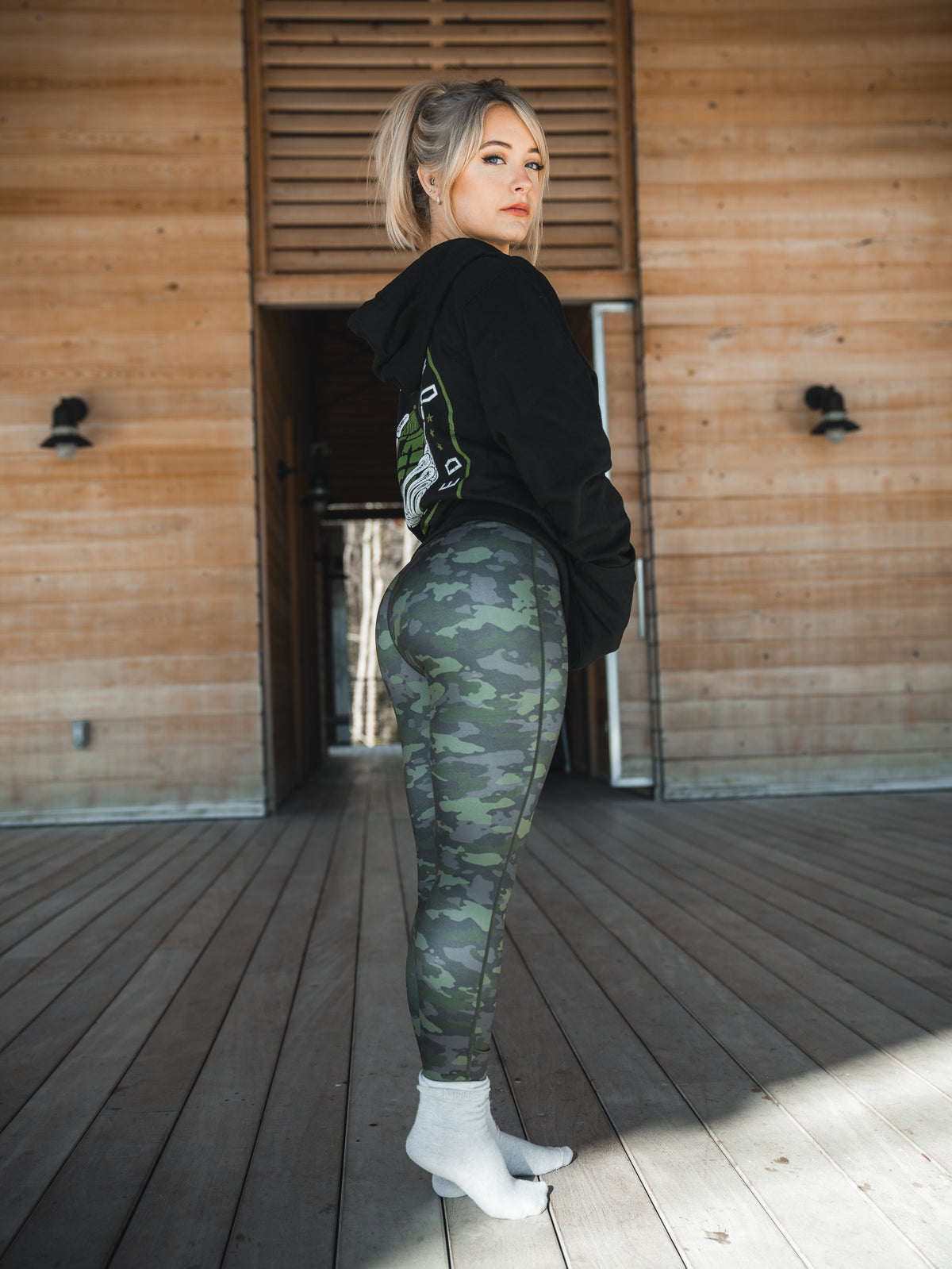 A woman stands on a wooden deck in front of a wooden building, wearing her favorite black hoodie and Born United Private Labels Combat Camo Leggings. Her hair is tied back as she looks to the side, white socks peeking out beneath her leggings.