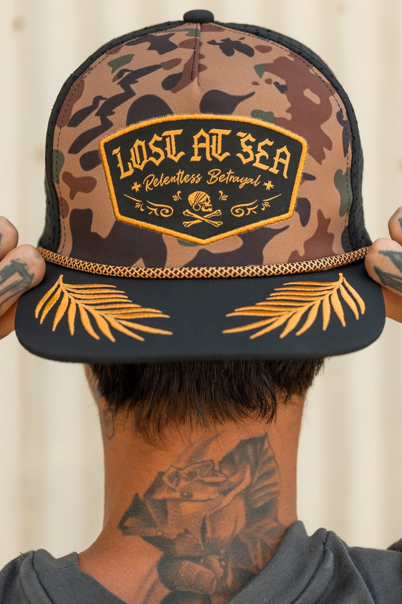 A tattooed person faces away wearing a Lost At Sea Frog Camo snapback by Relentless Betrayal, featuring a Lost at Sea, Relentless Betrayal patch with a skull design and a brim adorned with yellow palm motifs, giving an edgy vibe.