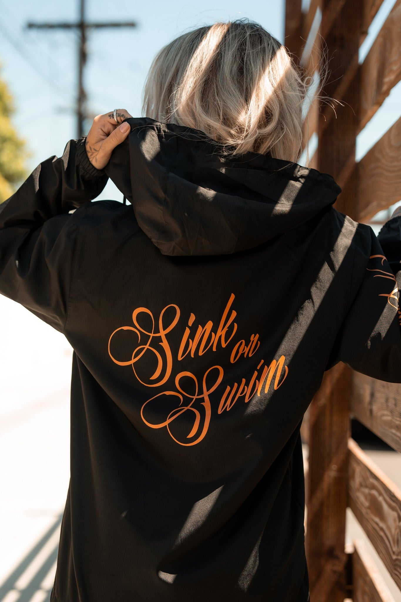 A blonde person stands outdoors in a Relentless Betrayal Sink or Swim Full Zip Jacket. The black windbreaker, with orange script on the back, glistens under the suns rays. A wooden fence and electrical pole are visible in the background.