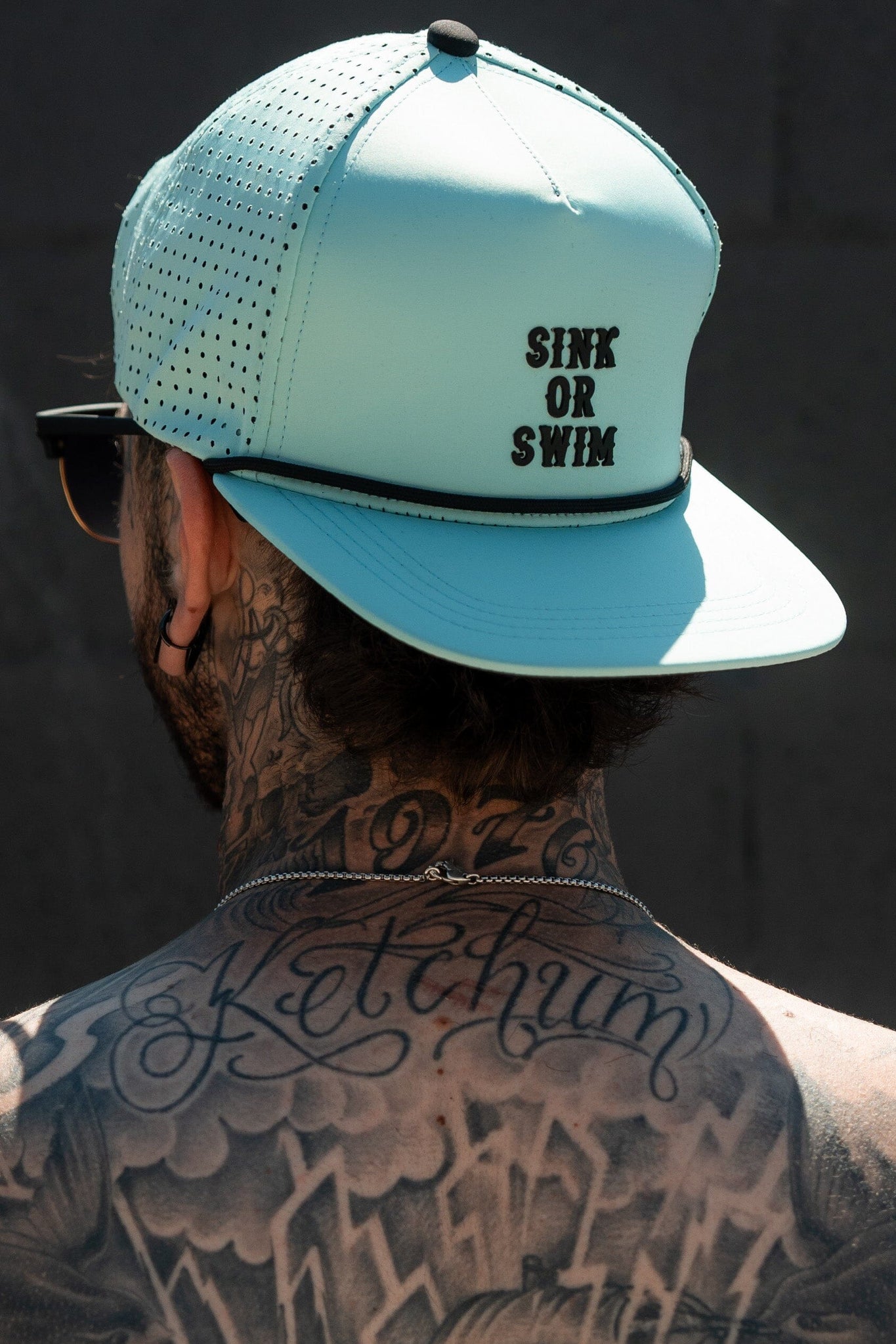 A person displays a tattooed neck and upper back while wearing the Sink or Swim Seafoam PVC Snapback by Relentless Betrayal, ideal for a pirate lifestyle. This water-resistant hat adds flair as they stand against a dark outdoor wall.
