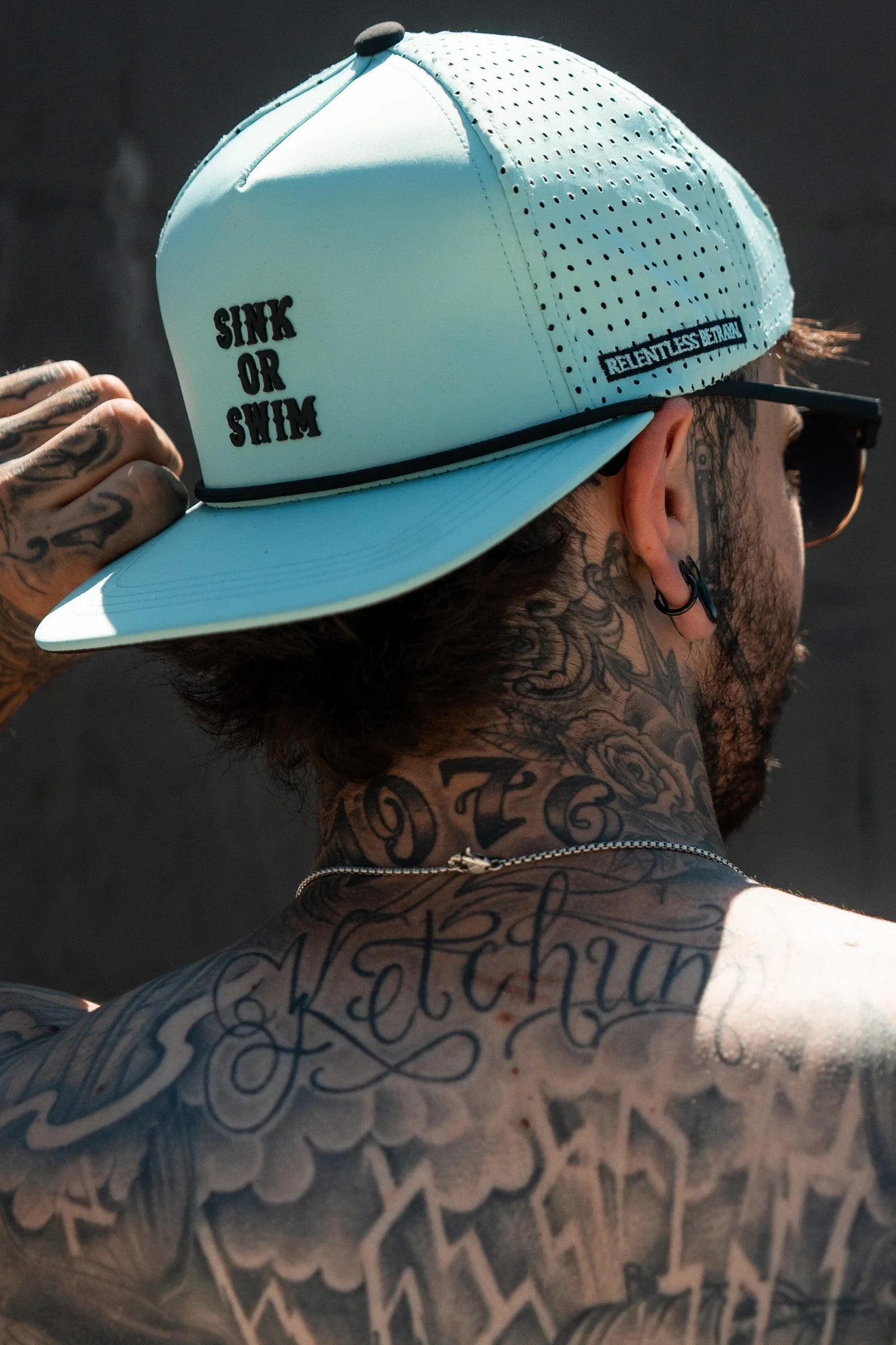 A tattooed man sports the Sink or Swim Seafoam PVC Snapback by Relentless Betrayal, featuring Sink or Swim on the front and Relentless on the side. Adjusting it, he embodies a pirate lifestyle, his back and neck tattoos fully visible from behind.