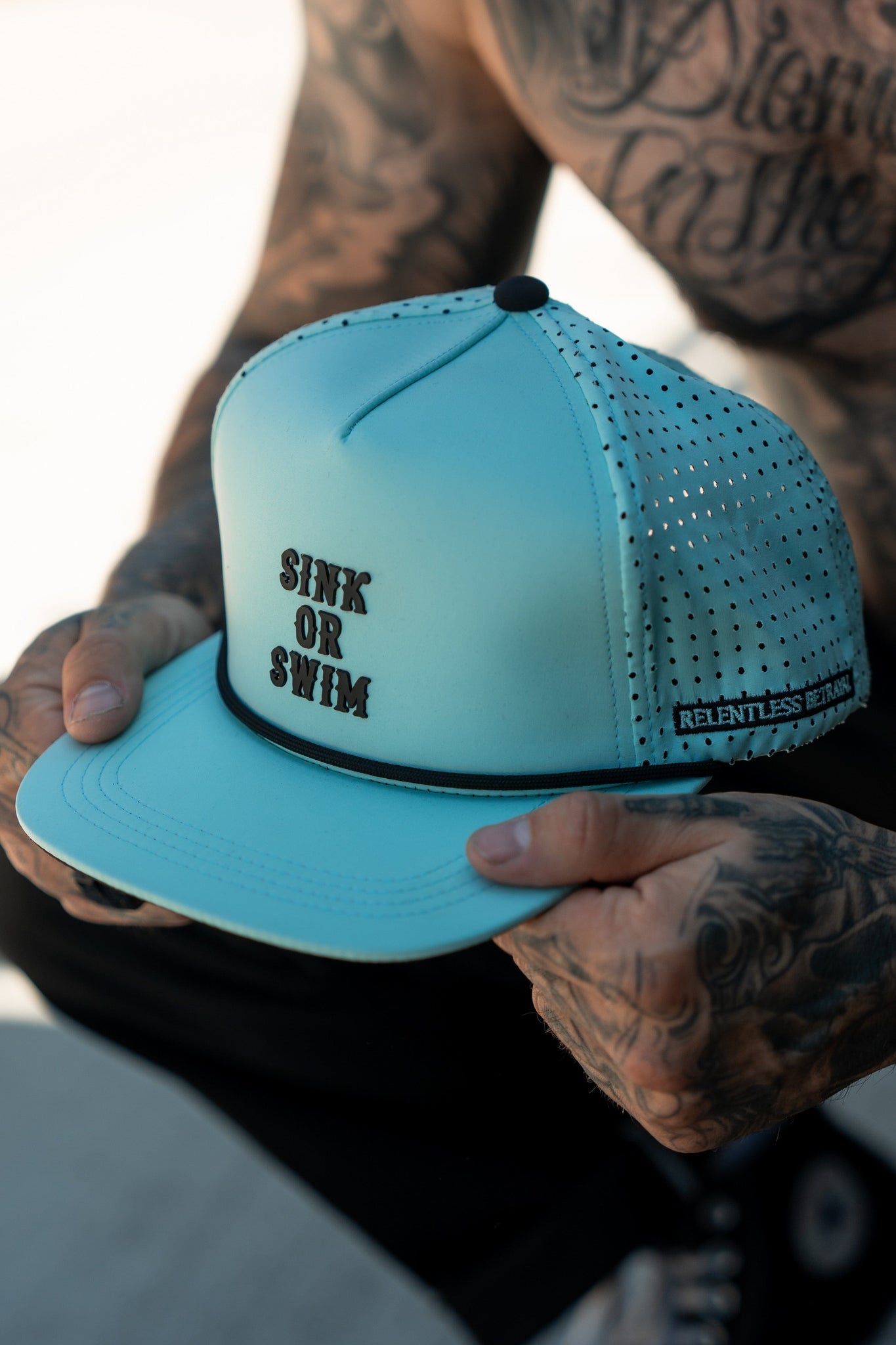Tattooed arms hold the Sink or Swim Seafoam PVC Snapback by Relentless Betrayal, featuring SINK OR SWIM on the front and RELENTLESS RETAIL on the side brim. The mesh sides embody its rugged, pirate spirit.
