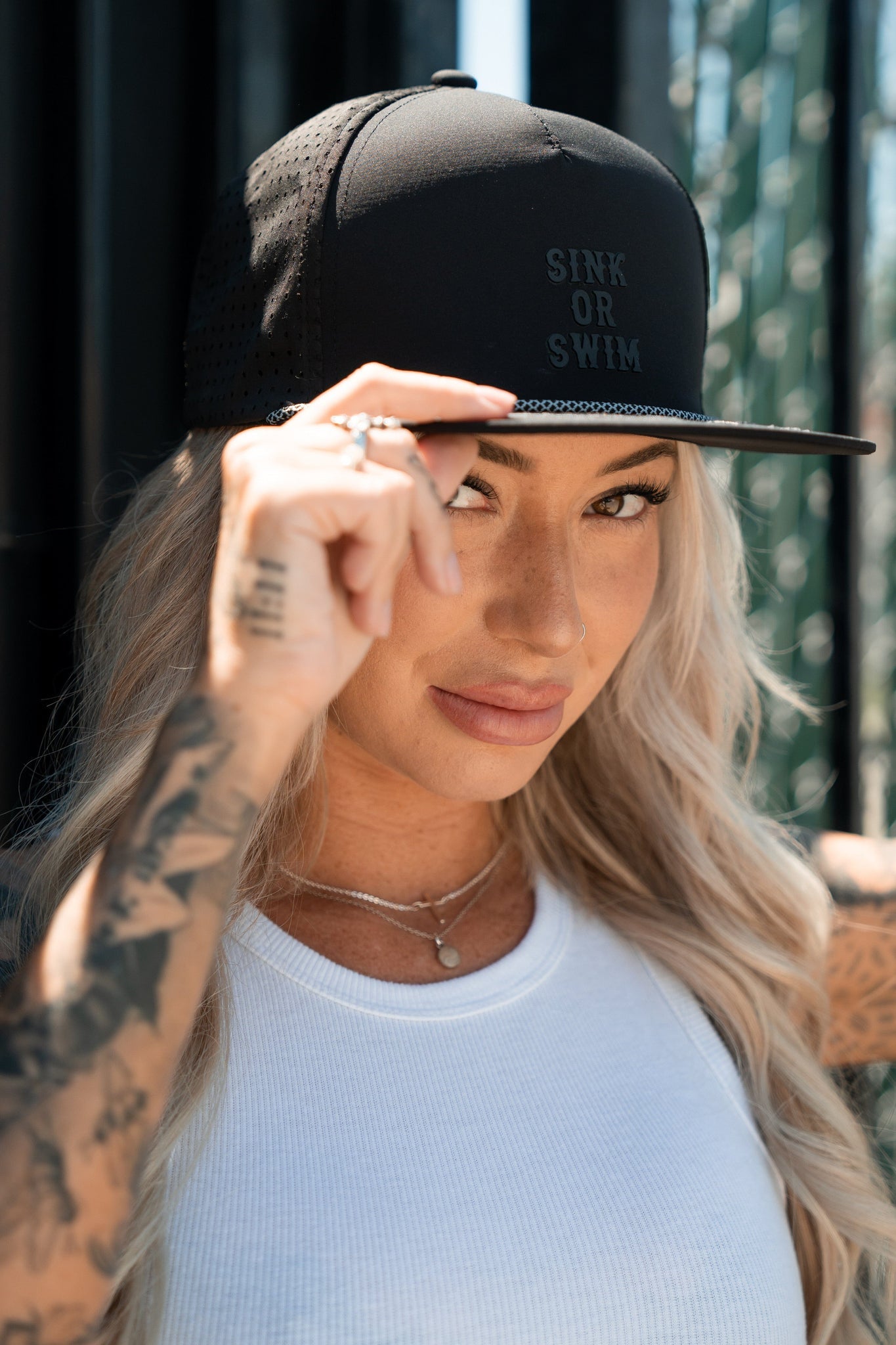 With long, wavy hair flowing under the Sink or Swim Blackout PVC Snapback by Relentless Betrayal, adorned with a 3D rubber patch, they embody a pirate lifestyle. Tattoos grace their arm as they hold the brim with one hand, smiling slightly while standing outdoors in their white top.