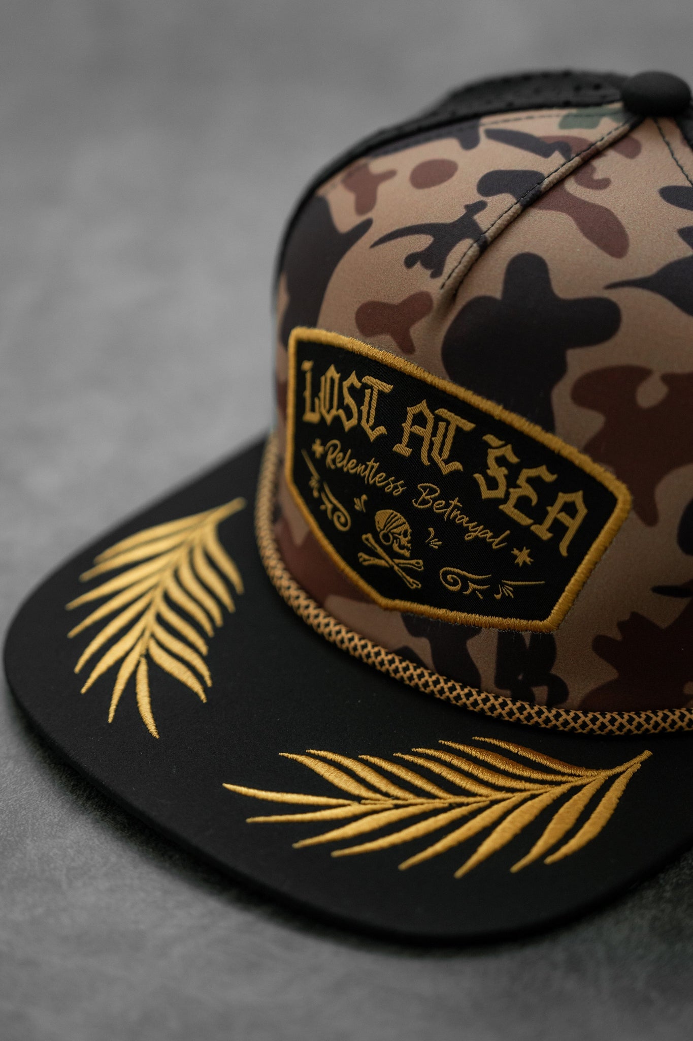 The Lost At Sea Frog Camo Snapback by Relentless Betrayal features a black and gold embroidered Lost At Sea patch on the front, water-resistant fabric, and intricate gold feather designs on the brim for a stylish yet practical look.