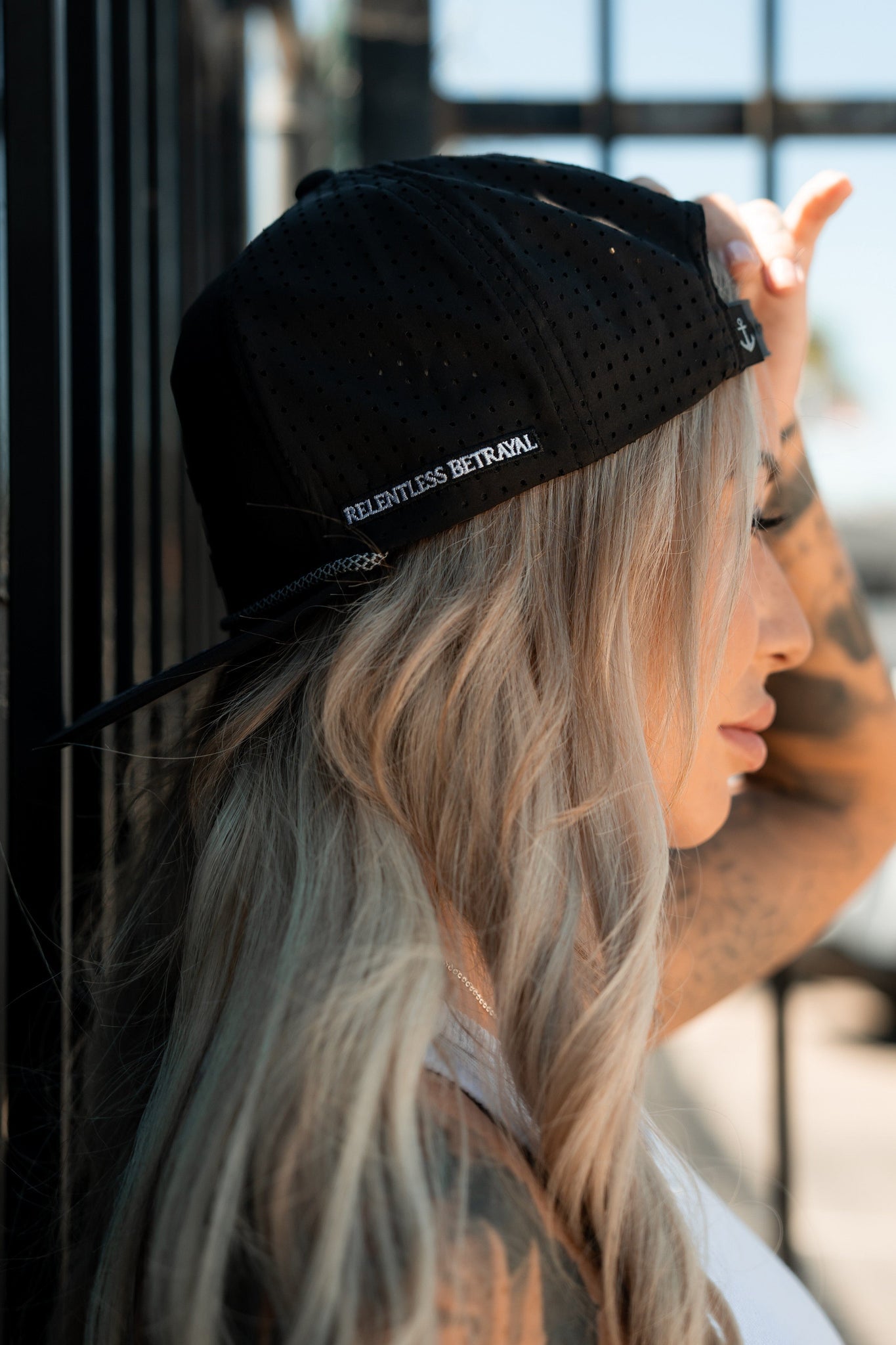 A person with long blonde hair, showcasing tattooed arms, wears the Sink or Swim Blackout PVC Snapback by Relentless Betrayal. The black water-resistant cap, turned backwards and adorned with a 3D PVC rubber patch of RELENTLESS BETRAYAL, stands out against a metal fence in the blurred background.