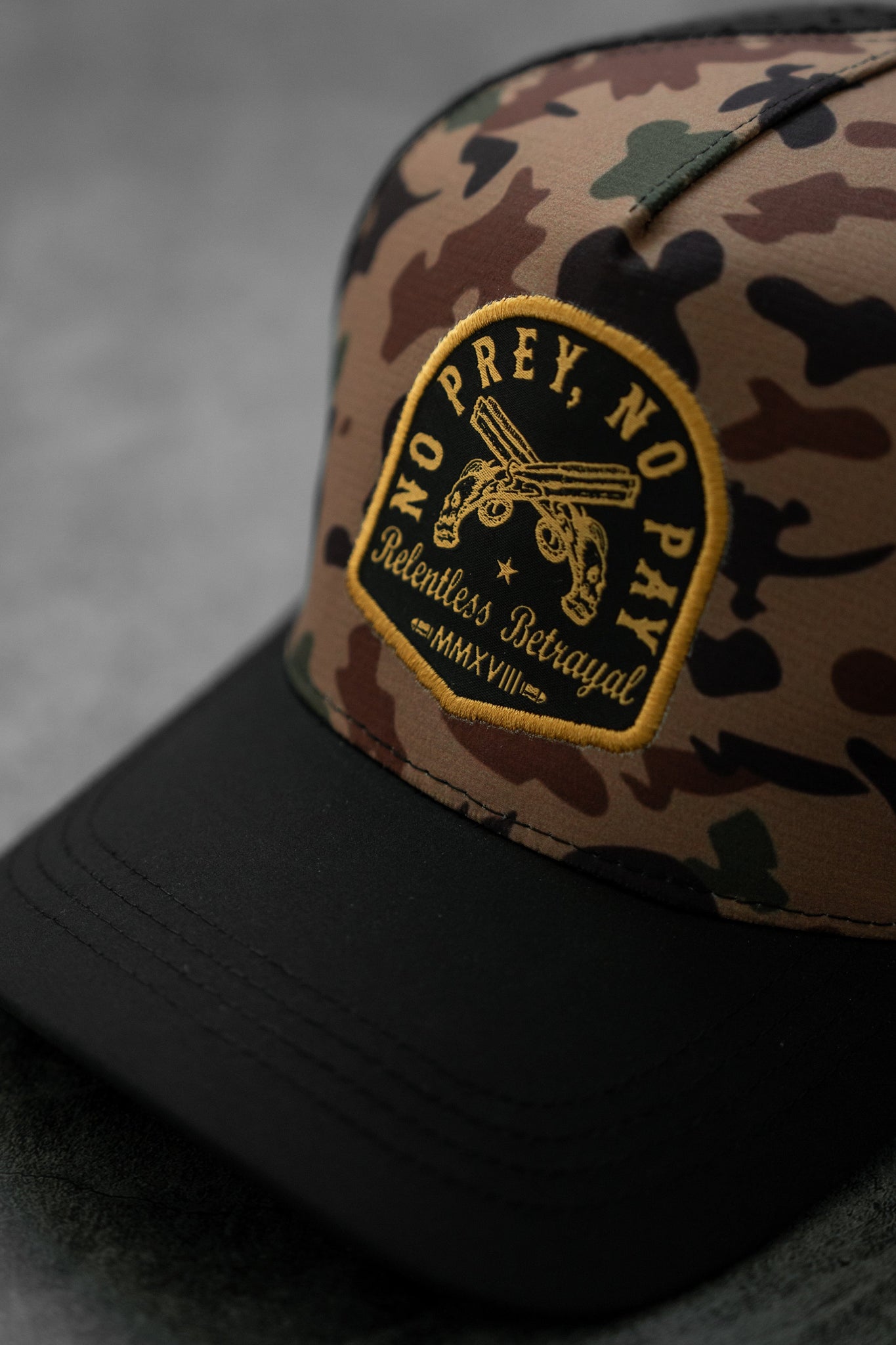 The No Prey, No Pay Frog Camo Hat by Relentless Betrayal is water-resistant, features a black brim, and displays crossed sabers with the phrases No Prey, No Pay and Relentless Betrayal MMXVIII, capturing the adventurous pirate spirit against a blurred gray background.