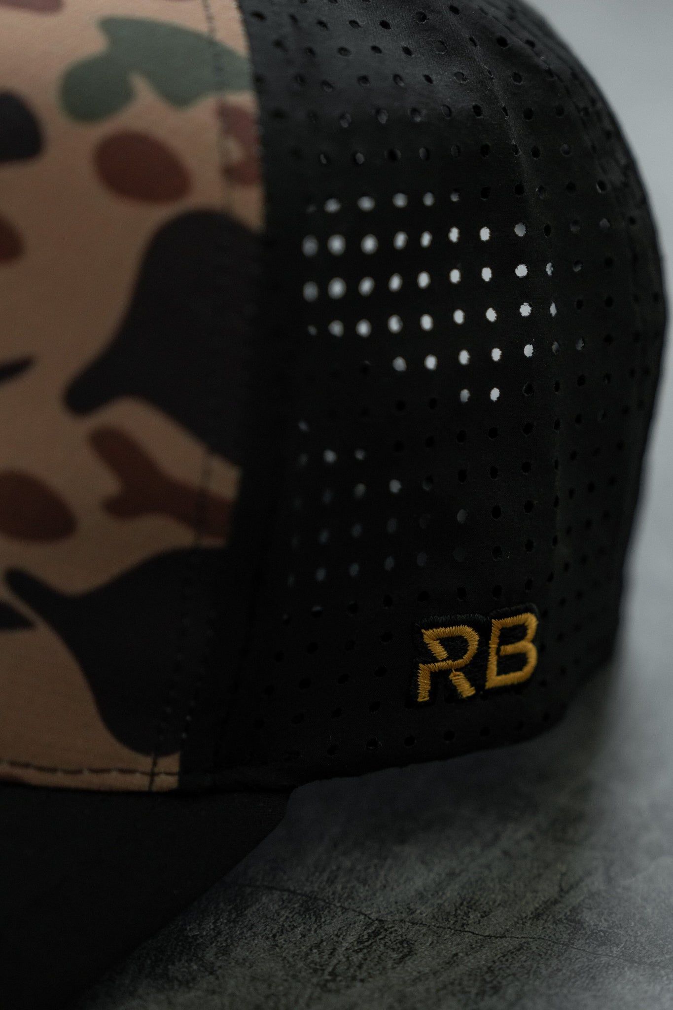 This close-up features the No Prey, No Pay Frog Camo Hat by Relentless Betrayal. It has a camo pattern on the front, water-resistant black mesh sides, and gold-embroidered RB initials for bold sophistication.