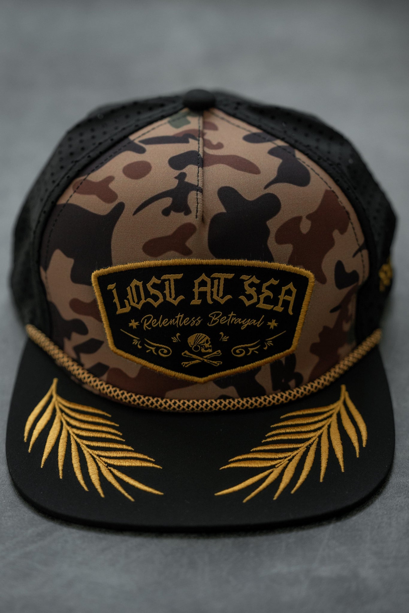 The Lost At Sea Frog Camo Snapback by Relentless Betrayal features a black front patch with Lost At Sea, Relentless Betrayal in vibrant yellow. Its brim has embroidered gold leaves, and its water-resistant design makes it ideal for any adventure.
