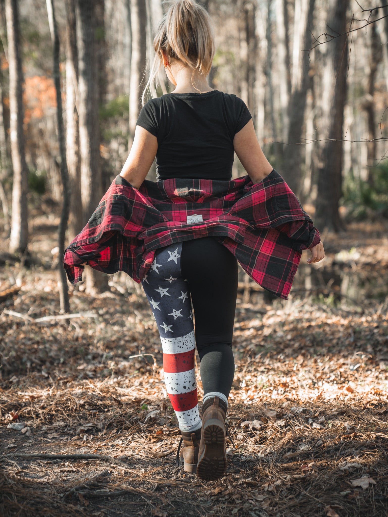 Strolling through the forest, a person with long hair sports a black shirt and Born United Private Labels American Savage Leggings, featuring stars and stripes on one leg. A red plaid jacket tied around their waist pairs perfectly with their brown boots as sunlight filters through the trees.