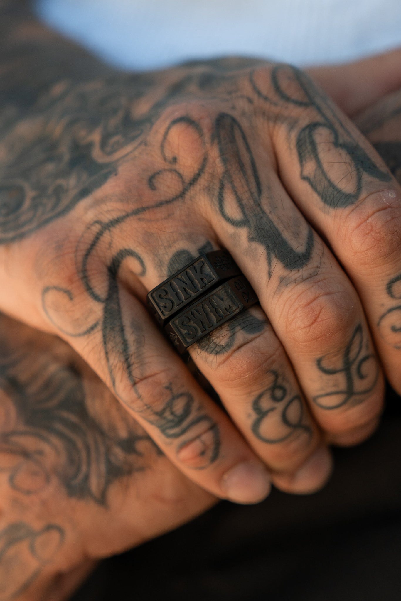 Close-up of a tattooed hand showcasing the Polished Black Sink or Swim Ring by Relentless Betrayal. The ornate tattoos complement the sleek design, and the warm skin tone contrasts with a blurred background, emphasizing this ocean-themed jewelry piece.