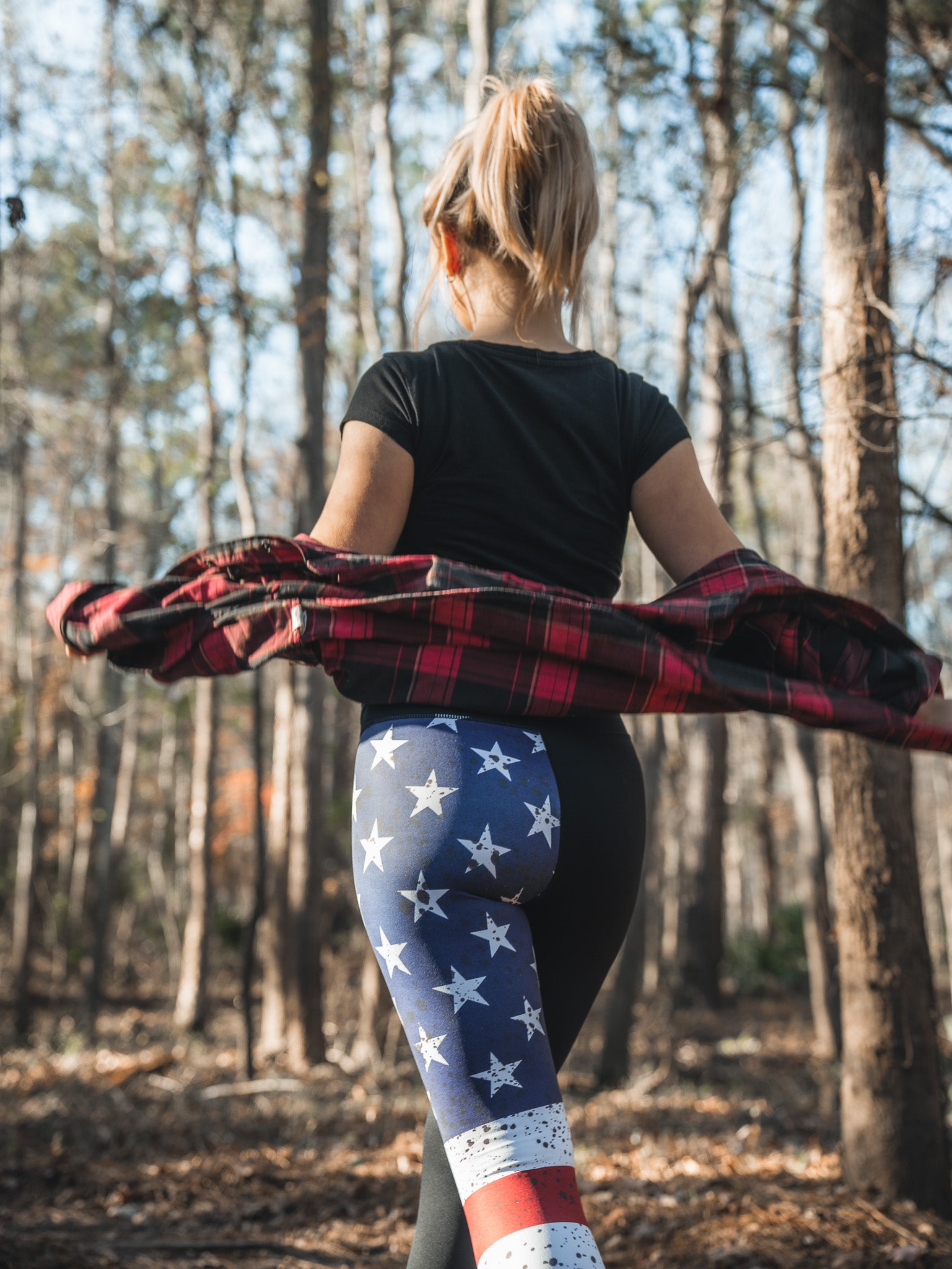 A woman strolls through the forest in American Savage Leggings by Born United Private Label, a black t-shirt, and clutching a red plaid shirt. Sunlight filters through the trees, creating a serene atmosphere.